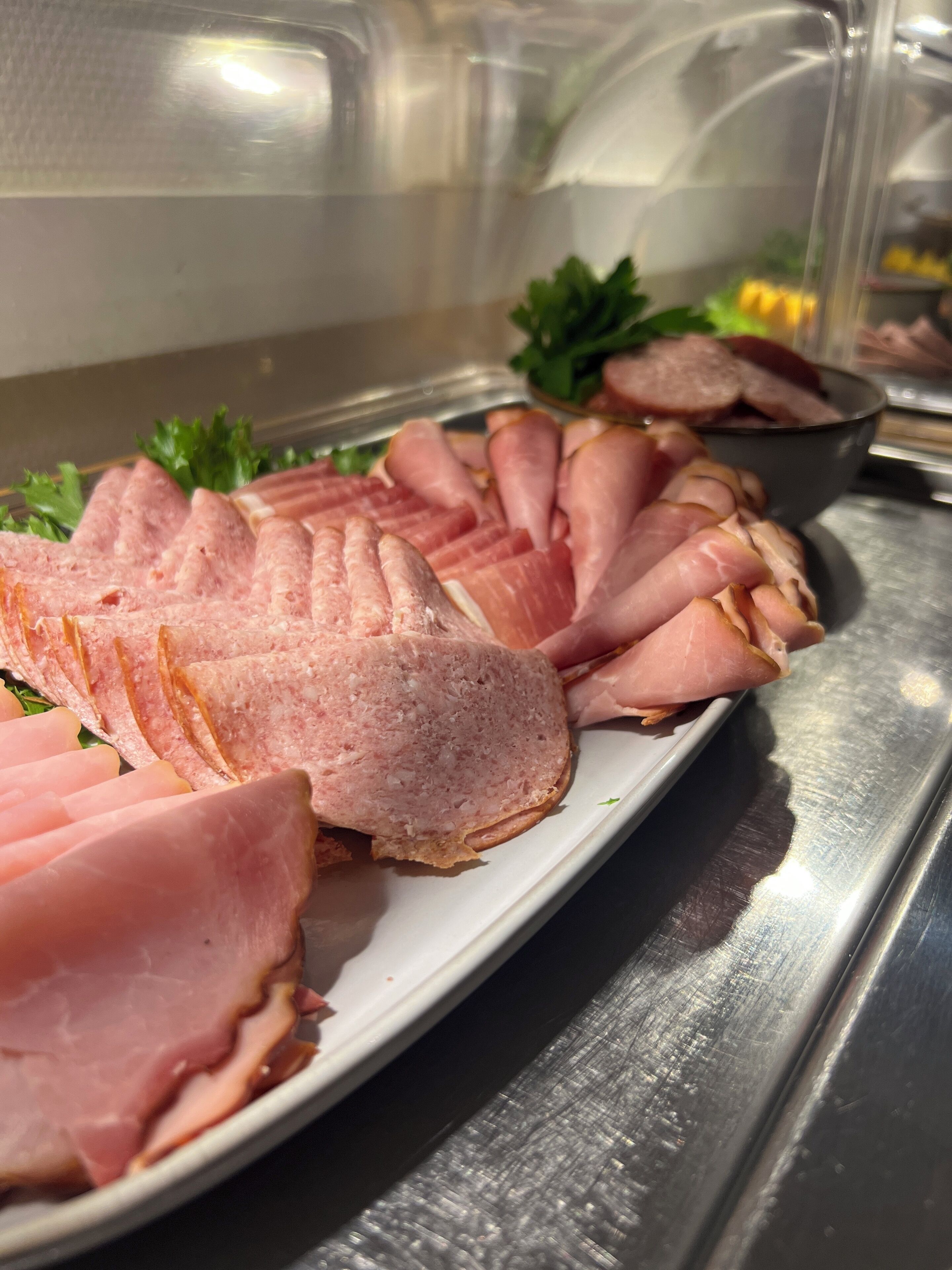 A display of sliced ham arranged on a white plate, with a metal counter and a glass enclosure in the background.