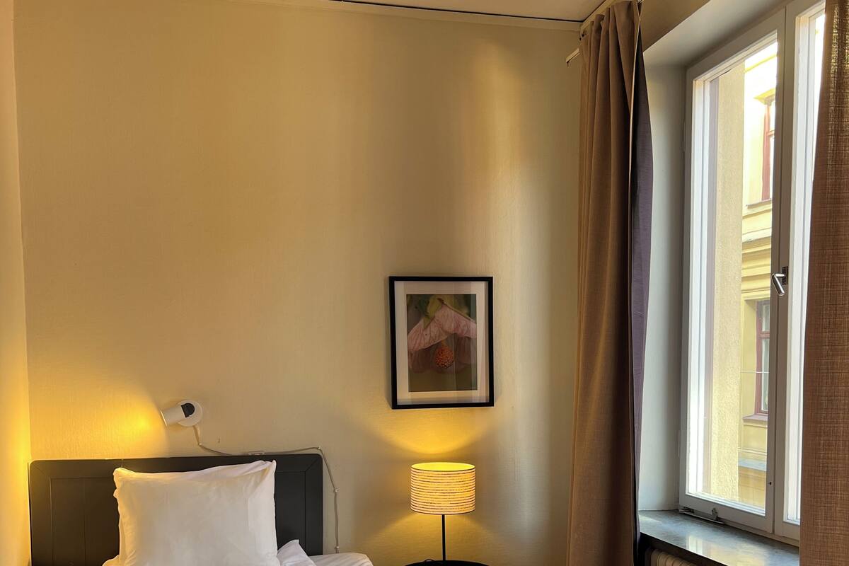 Quarto de hotel com cama, luminária de cabeceira, quadro na parede e janela com cortinas.