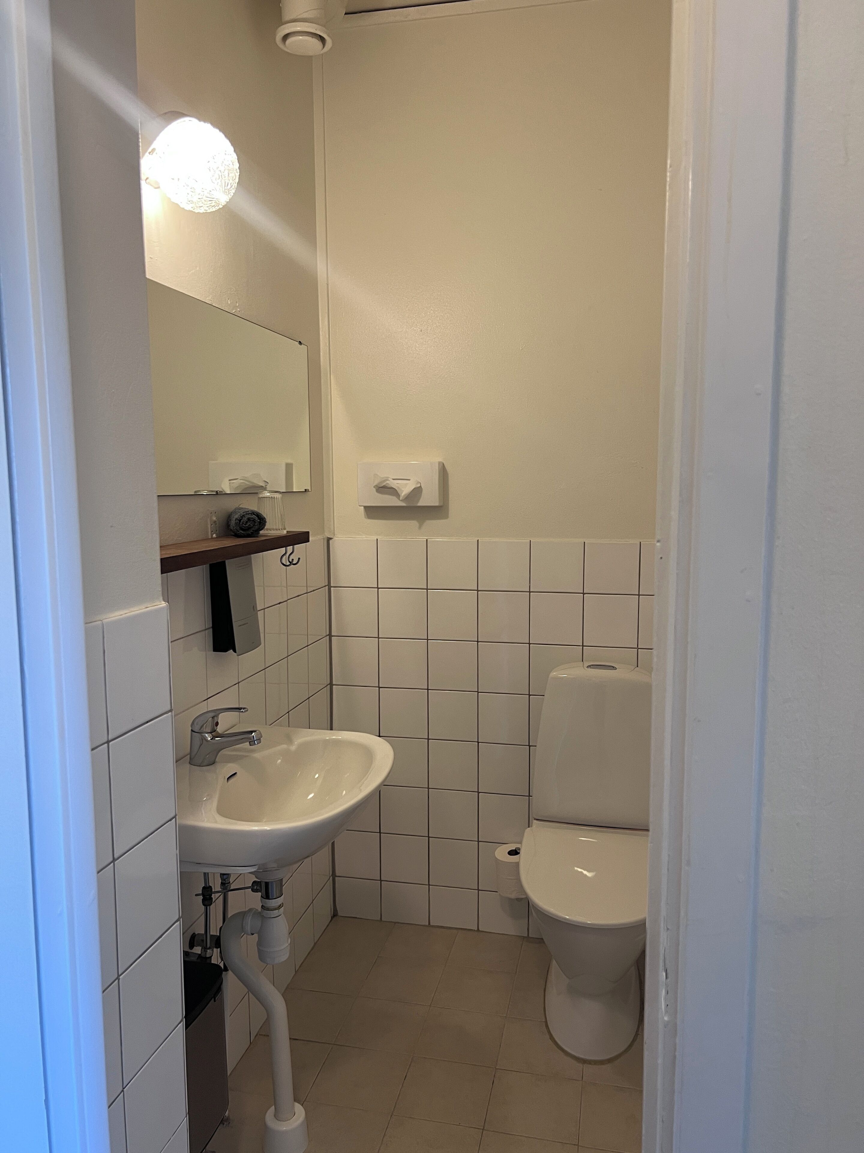 Une petite salle de bain propre, avec des carreaux blancs, un lavabo, des toilettes et un miroir.