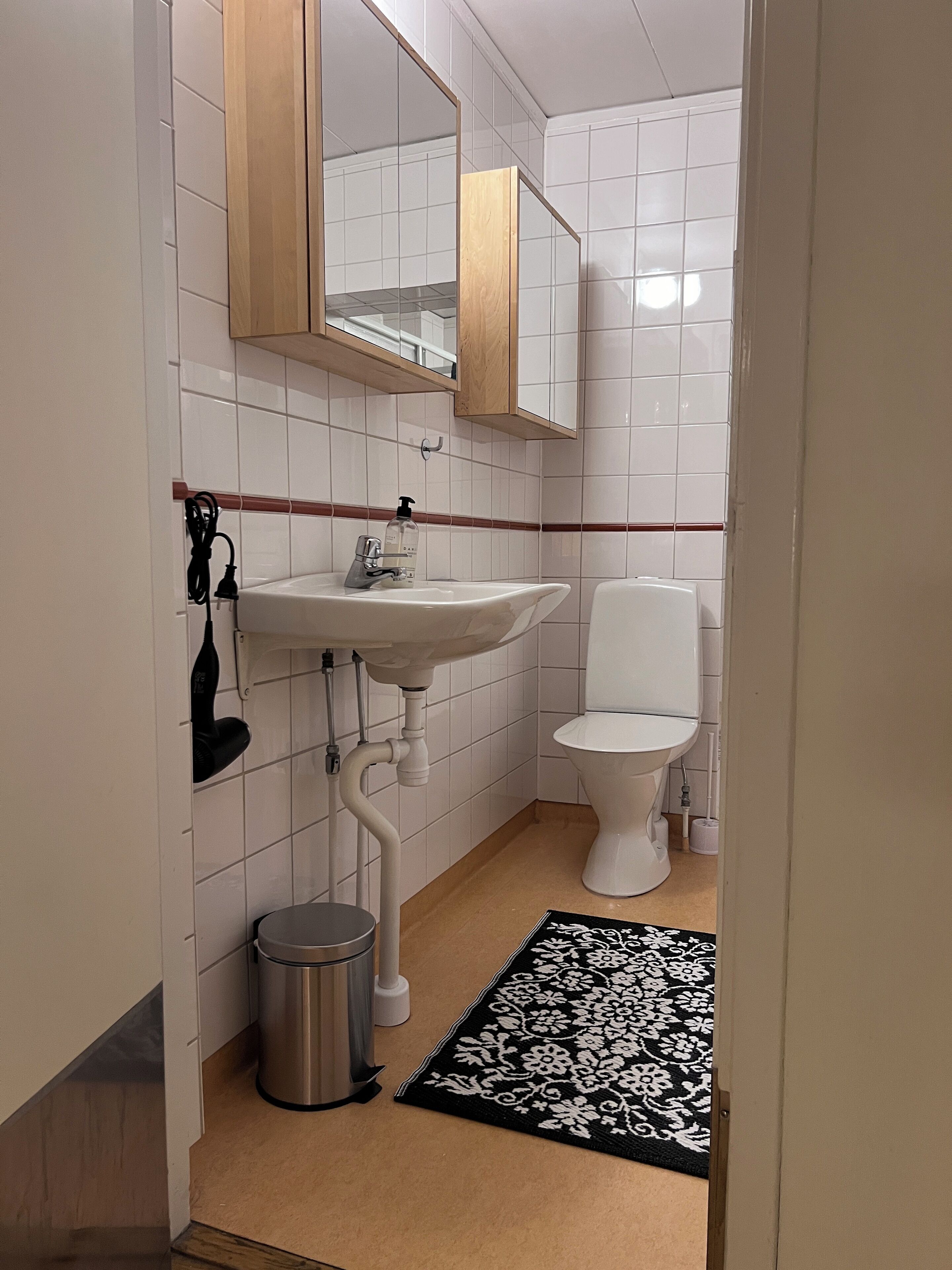 A small bathroom with white tiles, a sink, a toilet, a trash bin, and a black and white patterned rug.