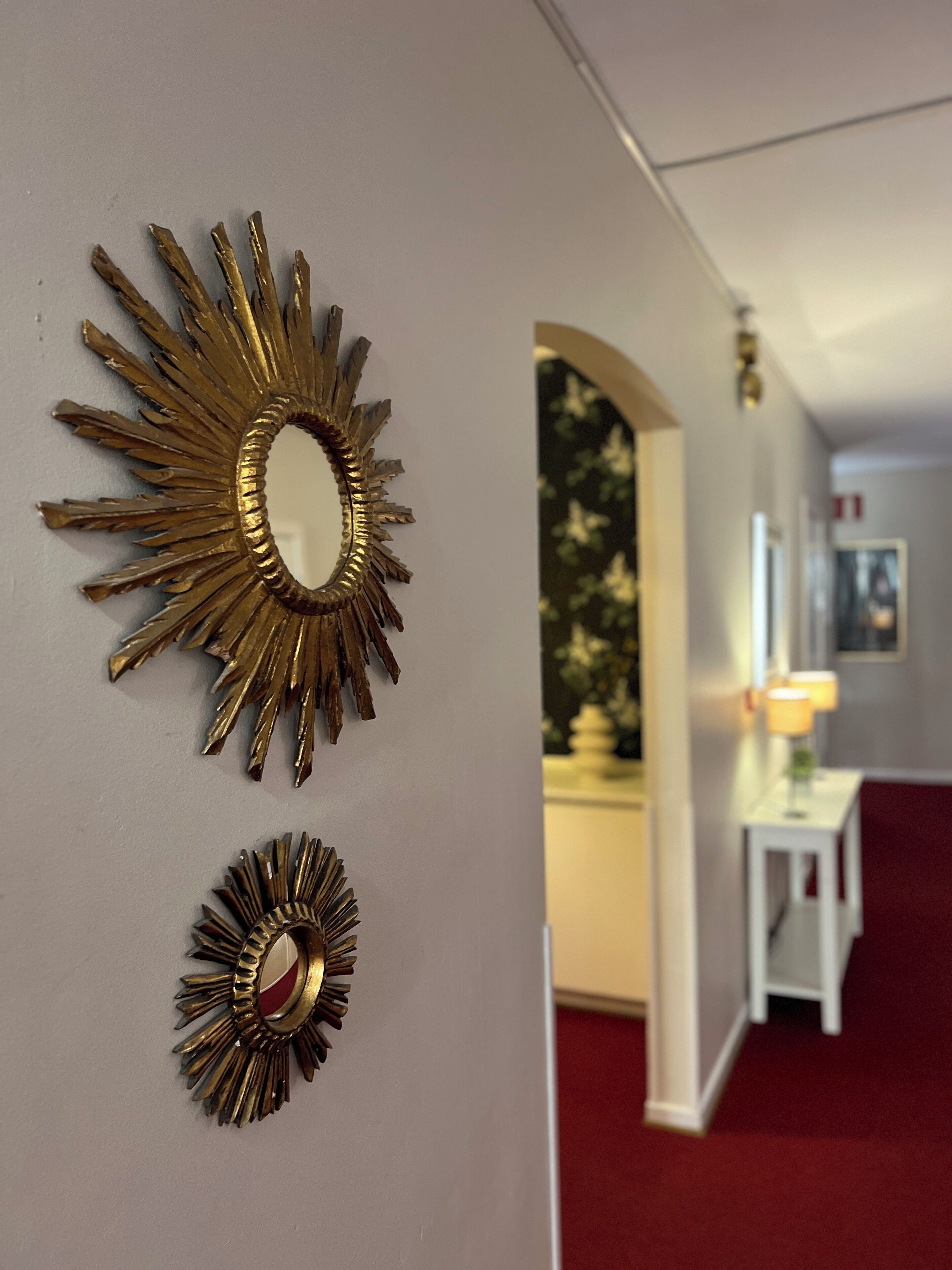 Un couloir orné de deux miroirs en forme de soleil sur le mur, une table blanche avec une lampe et un tapis rouge.