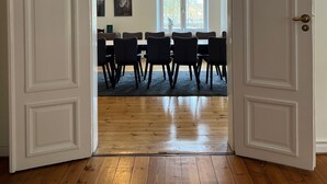 Une porte ouverte donne sur un espace repas doté d’un plancher en bois, d’une table entourée de chaises et d’appliques lumineuses suspendues au plafond.