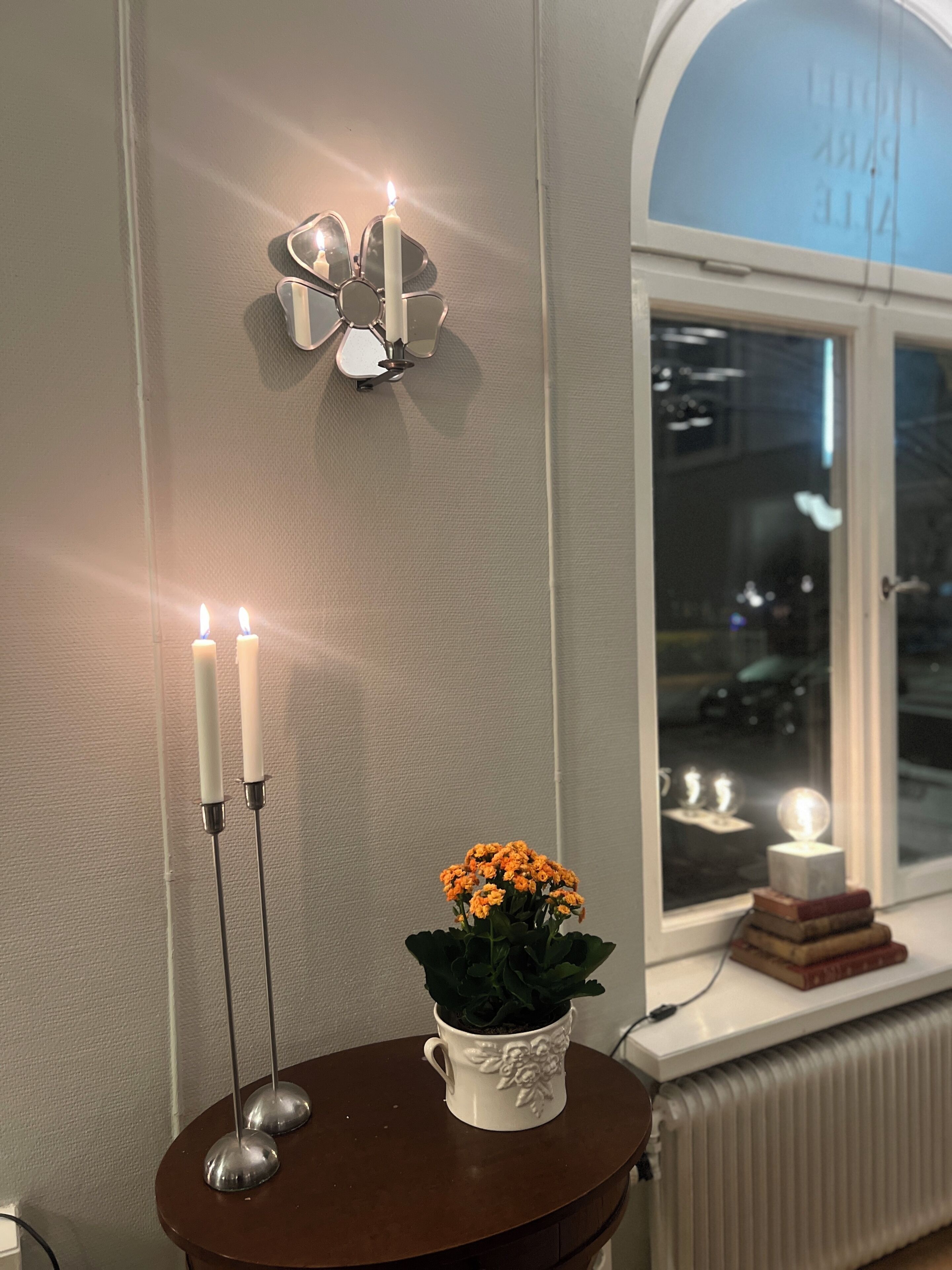 Una habitación con una lámpara de pared, dos velas, una planta en maceta y una ventana con vistas a la ciudad.