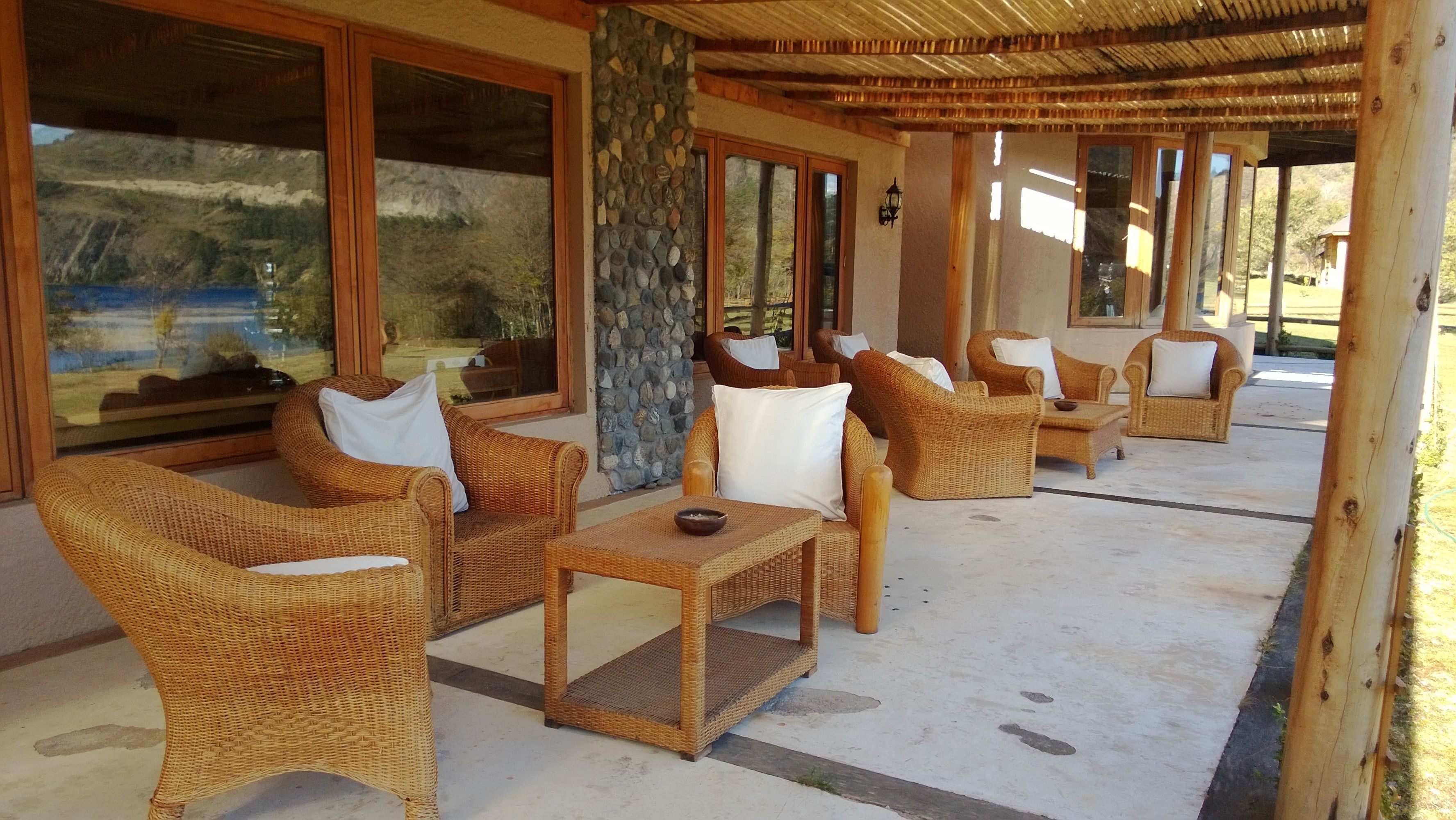 A covered outdoor seating area with wicker furniture and large windows overlooking a natural landscape.
