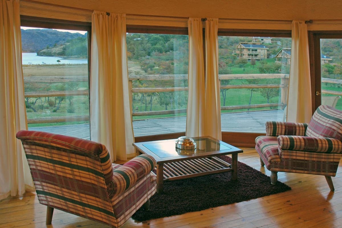Ein Zimmer mit zwei gestreiften Sesseln, einem kleinen Tisch und Blick auf einen See und Wald durch große Fenster.