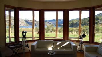 Habitación con ventana grande que ofrece vista a un paisaje montañoso y un lago. Hay un sofá blanco y un telescopio sobre un trípode.