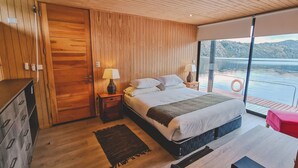 Un dormitorio moderno con techo de madera, una cama grande con ropa de cama blanca, una mesita de noche, una lámpara y vista a un lago a través de un ventanal grande.