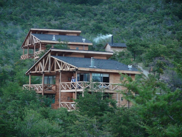 Una cabaña de madera de varios pisos con un balcón, rodeada por un bosque denso.