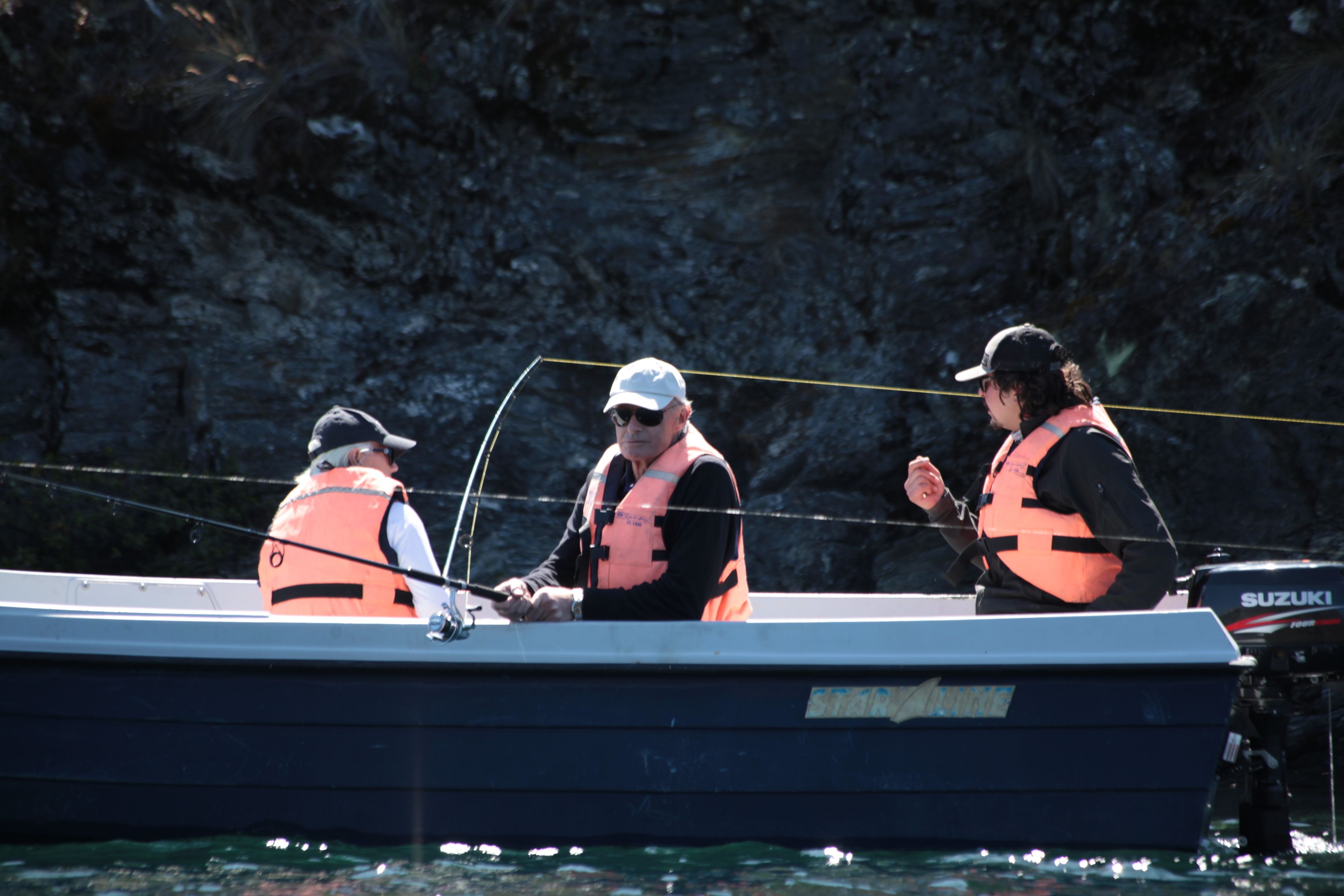 Three people on a boat with life jackets, a Suzuki outboard motor, and a rocky background.