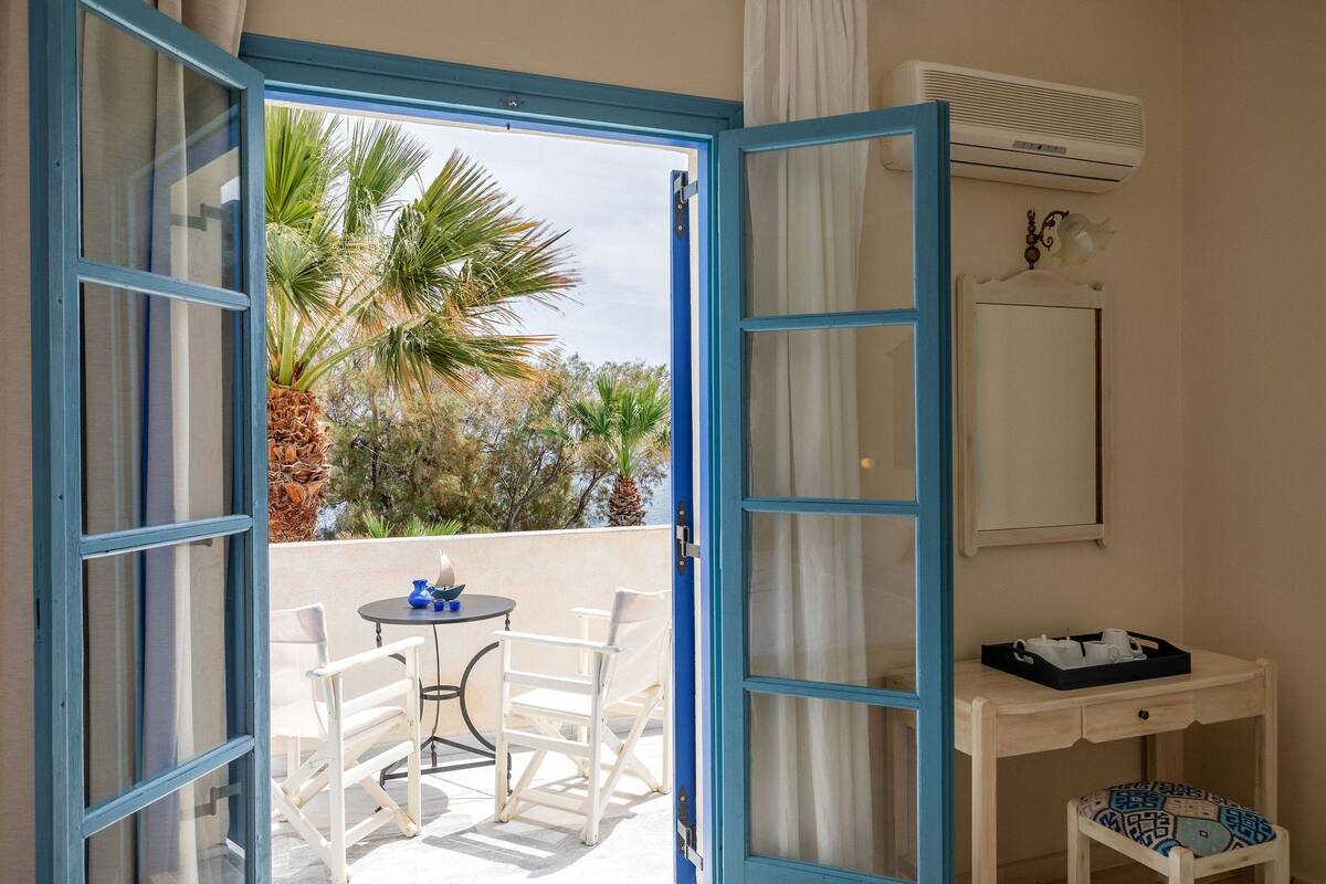 A room with a balcony, a table with chairs, a small desk with a chair, and an air conditioning unit.