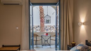 A hotel room with a bed, a small table with a red lamp, a chair, and a view of an outdoor area with a stone wall, a palm tree, and a hanging birdcage. - The Boathouse Hotel (Santorini)