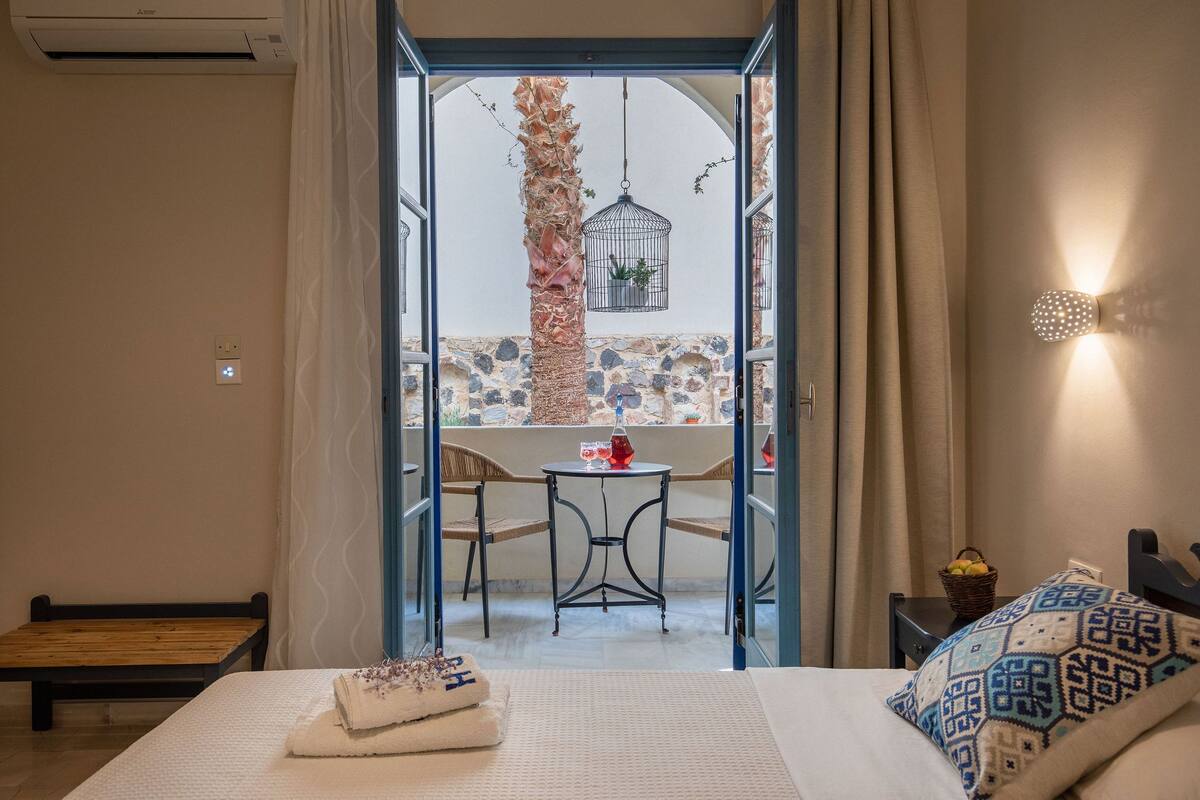 A hotel room with a bed, a small table with a red lamp, a chair, and a view of an outdoor area with a stone wall, a palm tree, and a hanging birdcage.