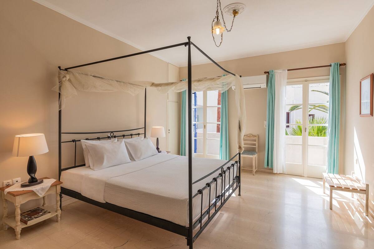 A four-poster bed with a canopy, bedside tables with lamps, a bench, and a view of the outdoors through a glass door.