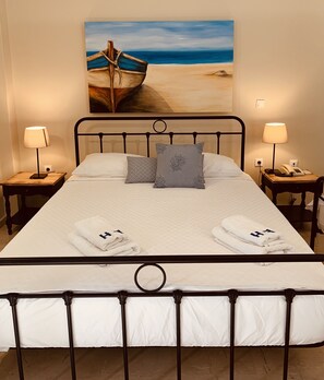 A neatly made bed with white linens, two pillows, and two folded towels. There are two bedside tables with lamps and a phone. A painting of a boat on a beach hangs on the wall above the bed. - The Boathouse Hotel (Santorini)