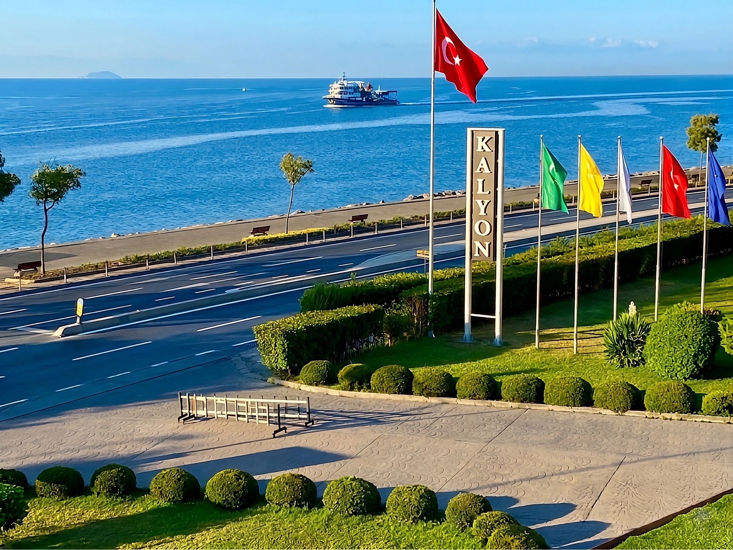 A coastal road with a sign reading 'KAYLON,' flags, and a body of water with a boat.