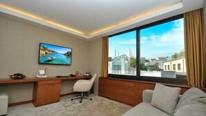 A hotel room with a large window, a desk with a chair, a sofa, and a painting of a coastal scene. - Kalyon Hotel Istanbul (Istanbul)