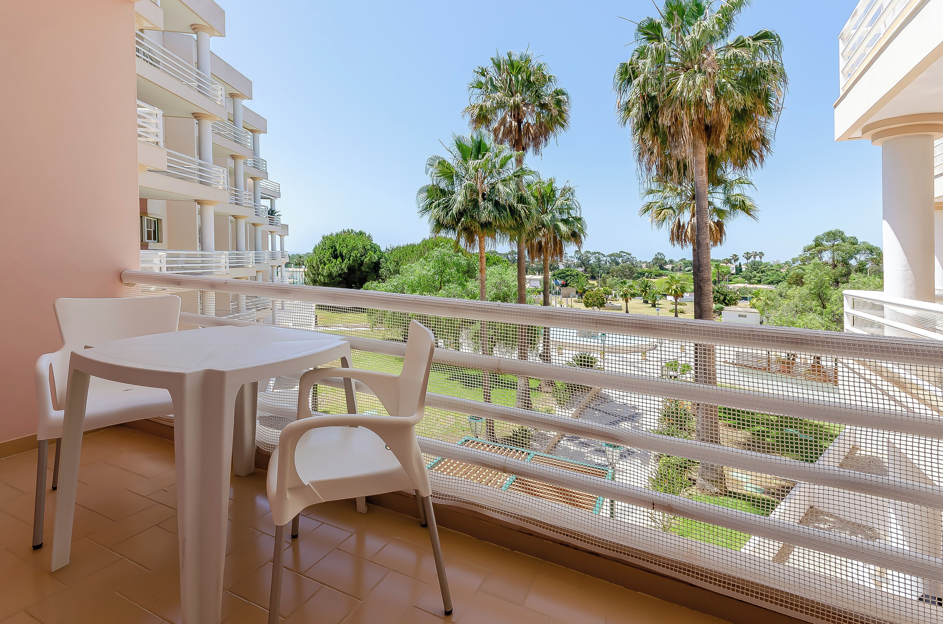 Varanda com mesa e cadeiras brancas, com vista para um jardim com palmeiras e um prédio ao fundo.