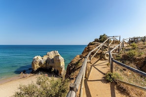 A coastal path with wooden railings leading to a beach, surrounded by rocky cliffs and clear blue sea.