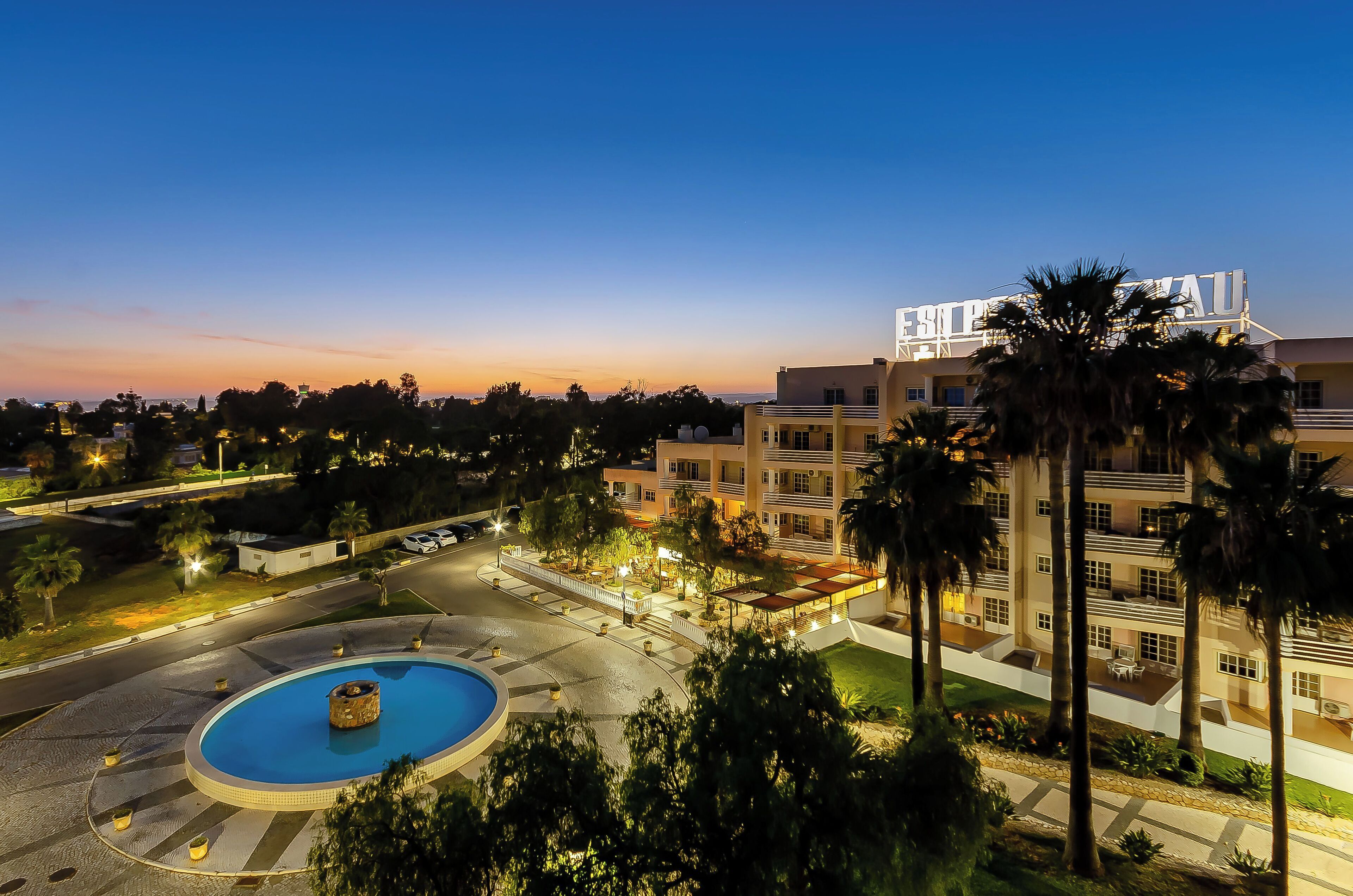 Un hotel con una piscina grande, palmeras y un patio bien iluminado.