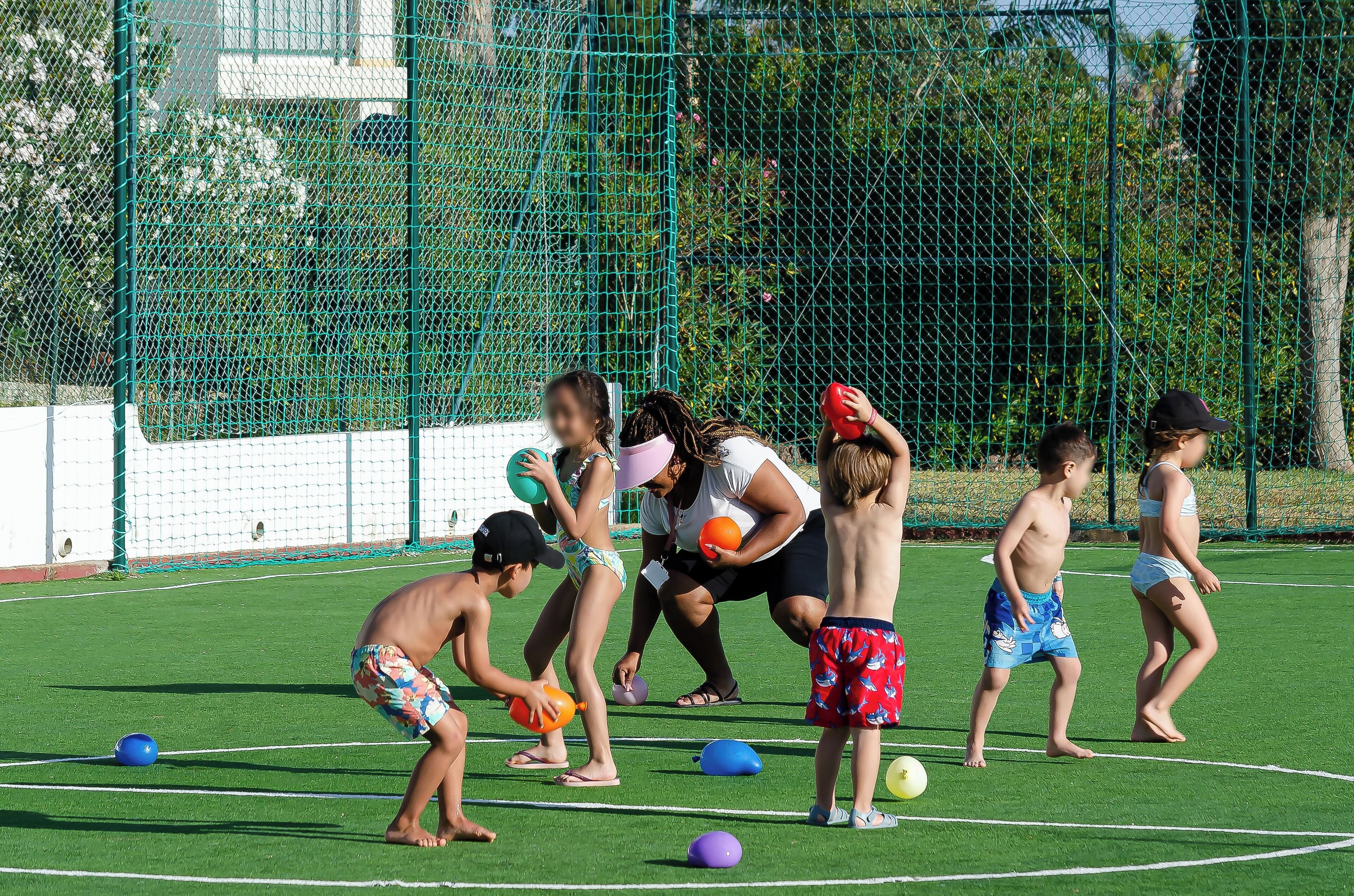 Children playing with balls on a green field with a fence in the background.