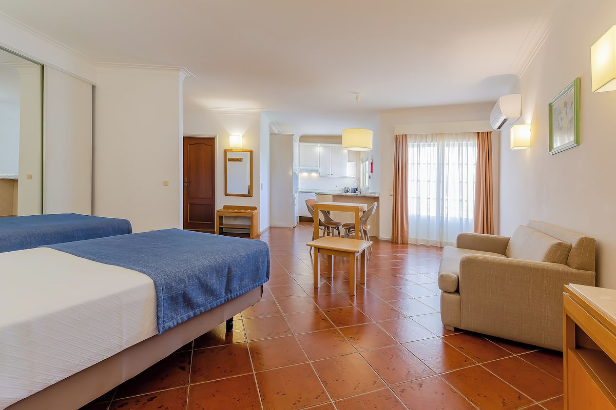 Een hotelkamer met twee bedden, een bank, een eettafel met stoelen en een kitchenette op de achtergrond.