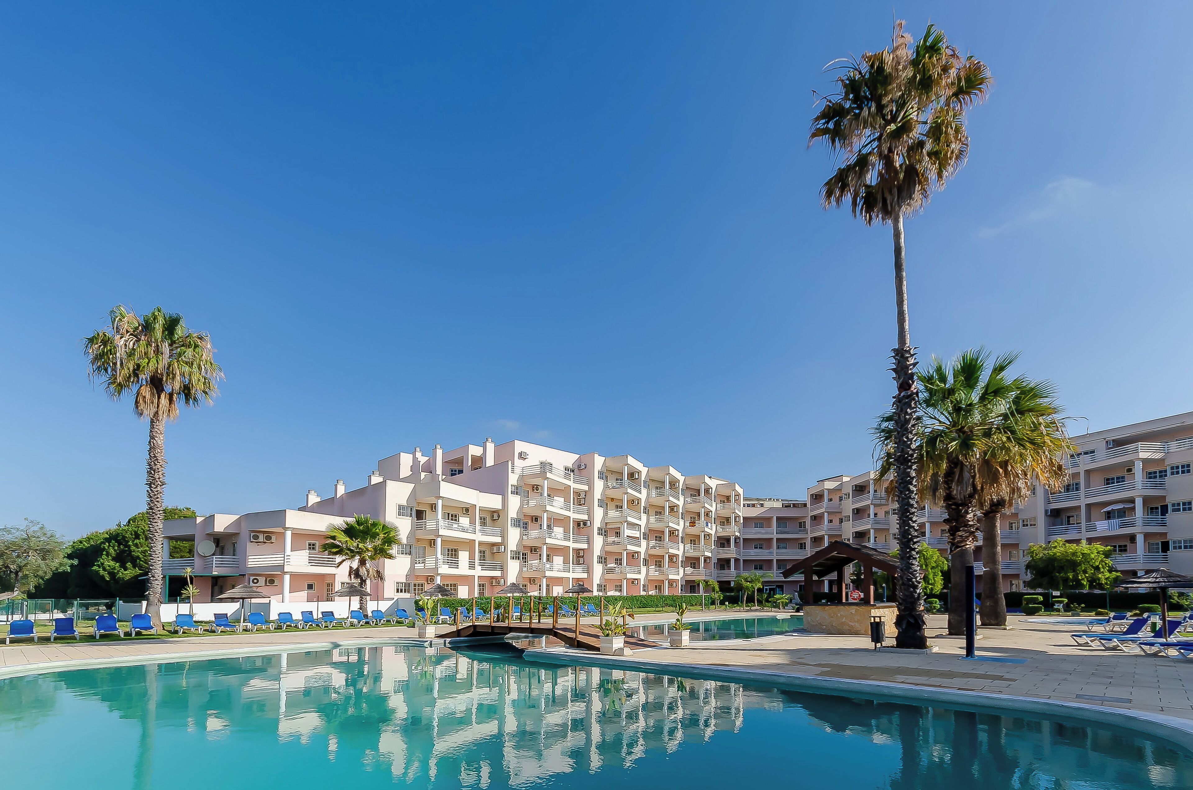 A hotel with a swimming pool, palm trees, and a clear blue sky.