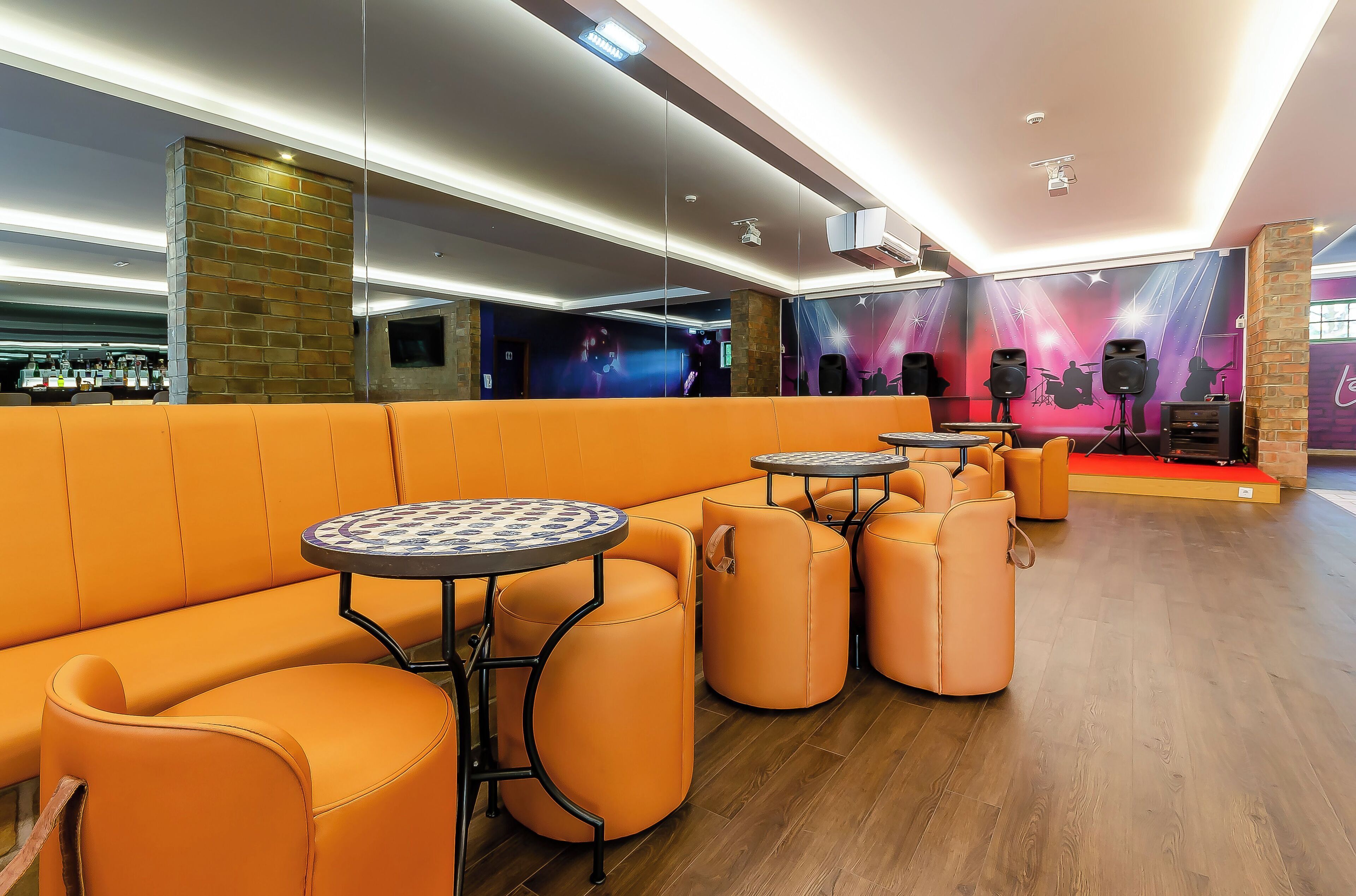 Un espacio lounge moderno con asientos anaranjados, mesas redondas y un escenario con instrumentos musicales.