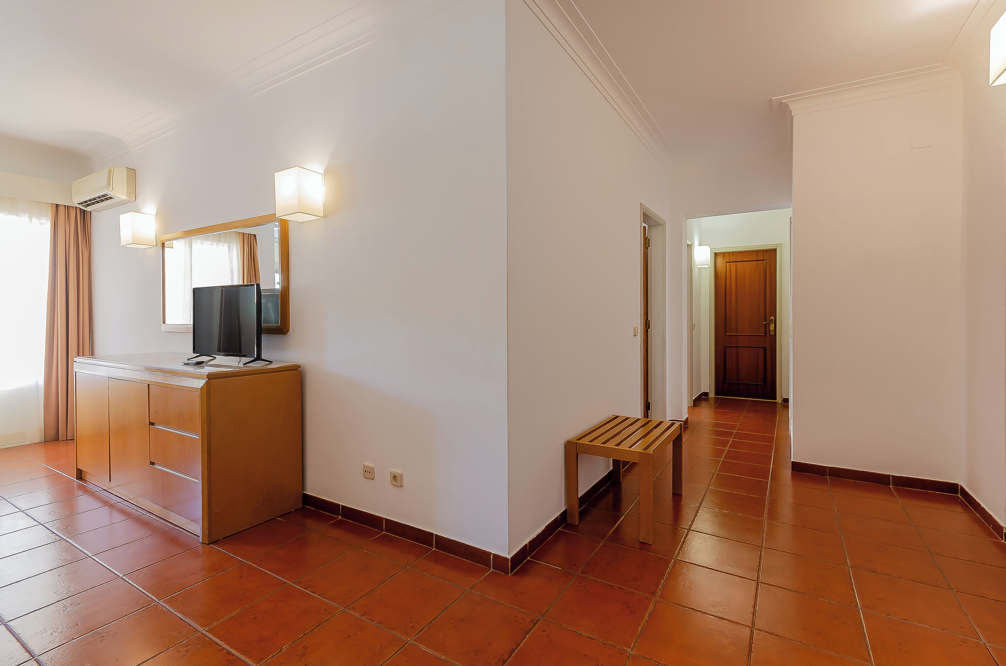 A hotel room with a minibar, a television, a mirror, and a hallway leading to a door.