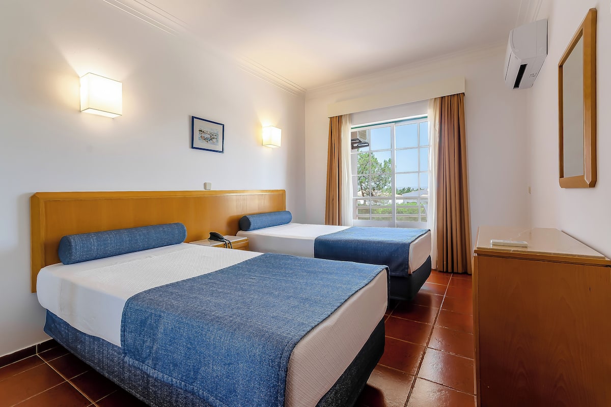 Hotelkamer met twee bedden, een houten hoofdeinde, een bureau, een stoel, een raam met gordijnen en een wandairconditioner.