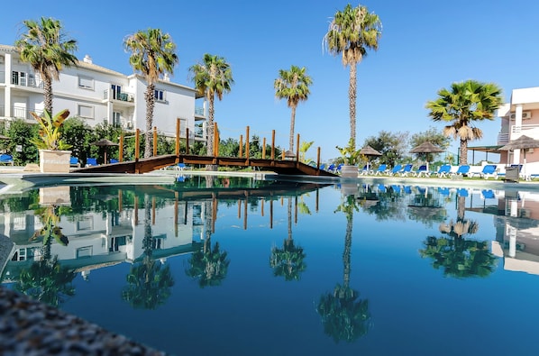 A pool area with clear reflections of palm trees and a wooden bridge.