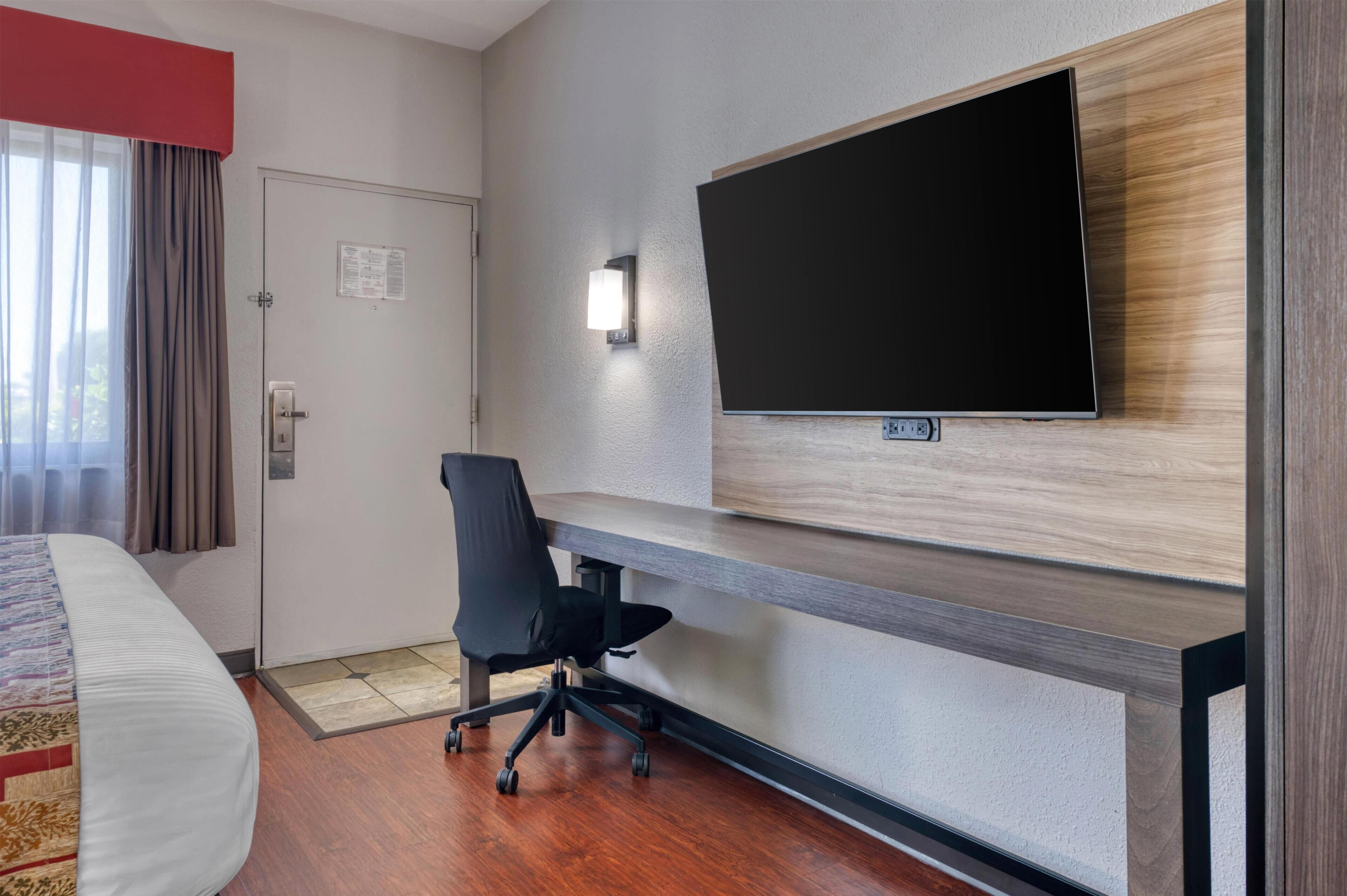 A hotel room with a bed, a desk, a chair, a TV mounted on the wall, and a door.