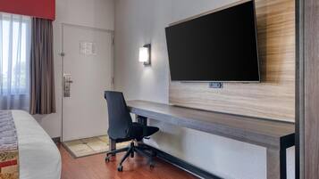 A hotel room with a bed, a desk, a chair, a TV mounted on the wall, and a door.