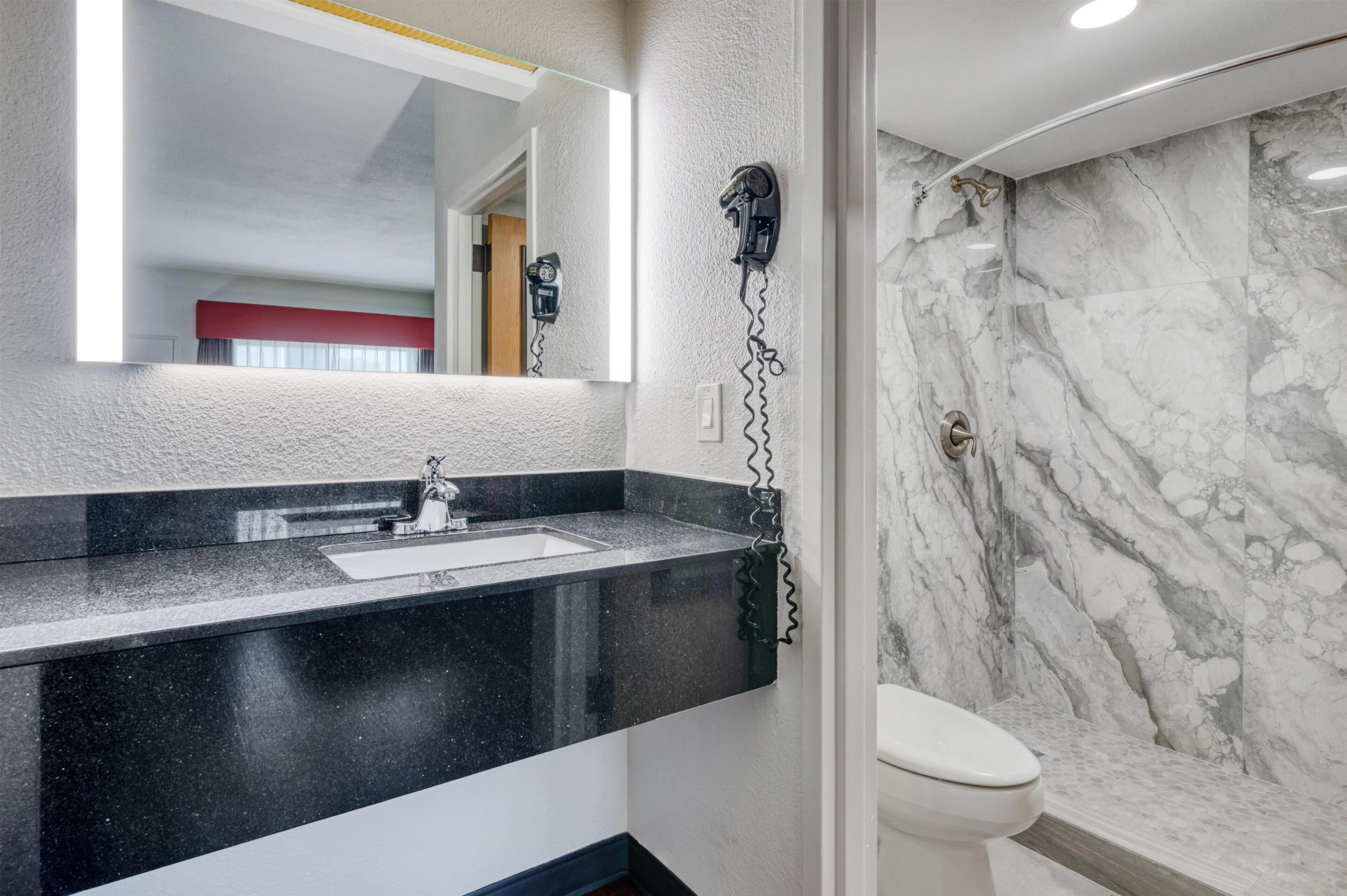 Un baño con encimera negra, lavamanos blanco, secador de pelo montado en la pared y pared de ducha de mármol.