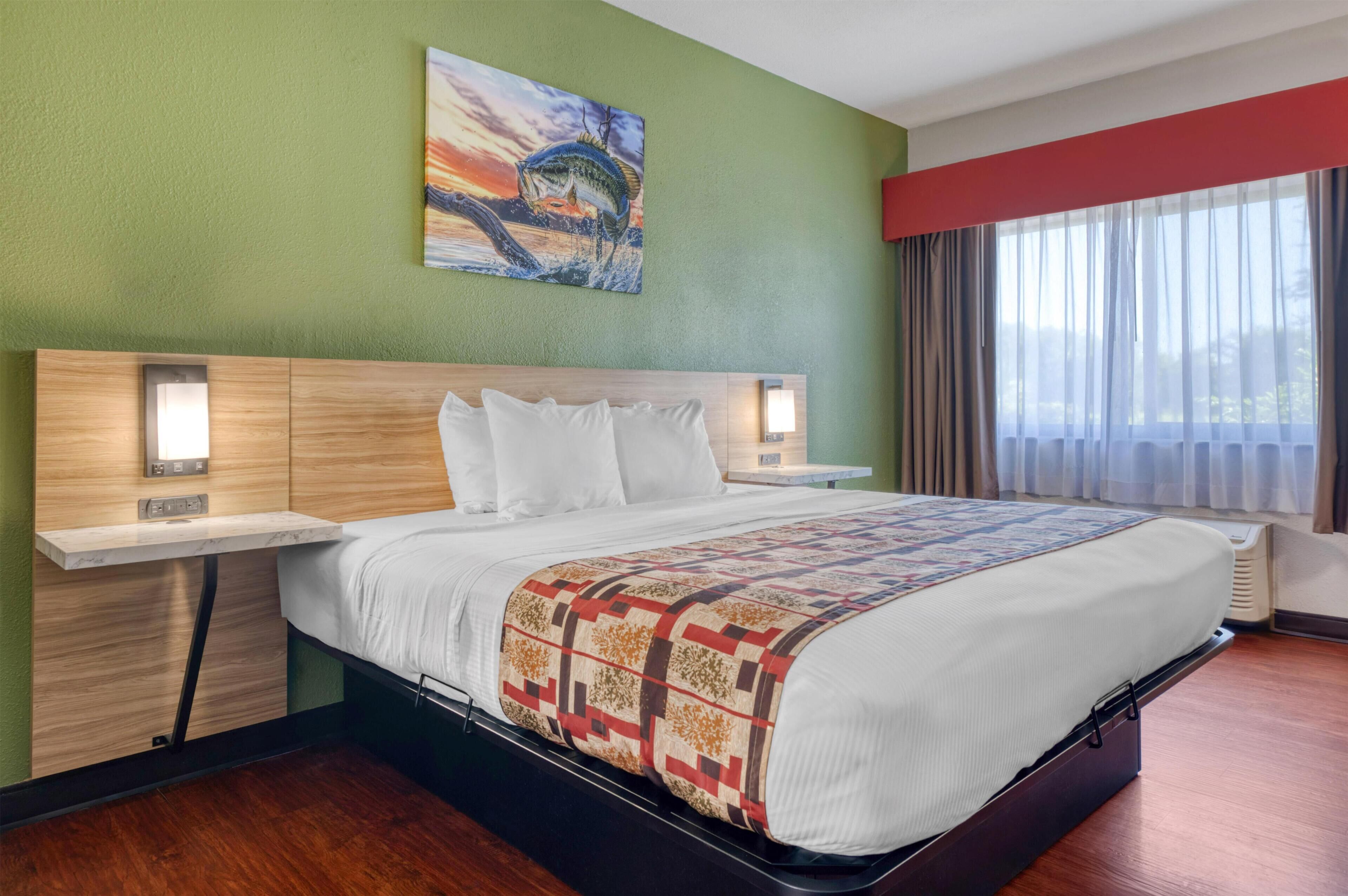 A hotel room with a large bed, a nightstand, a wall-mounted painting, and a window with curtains.