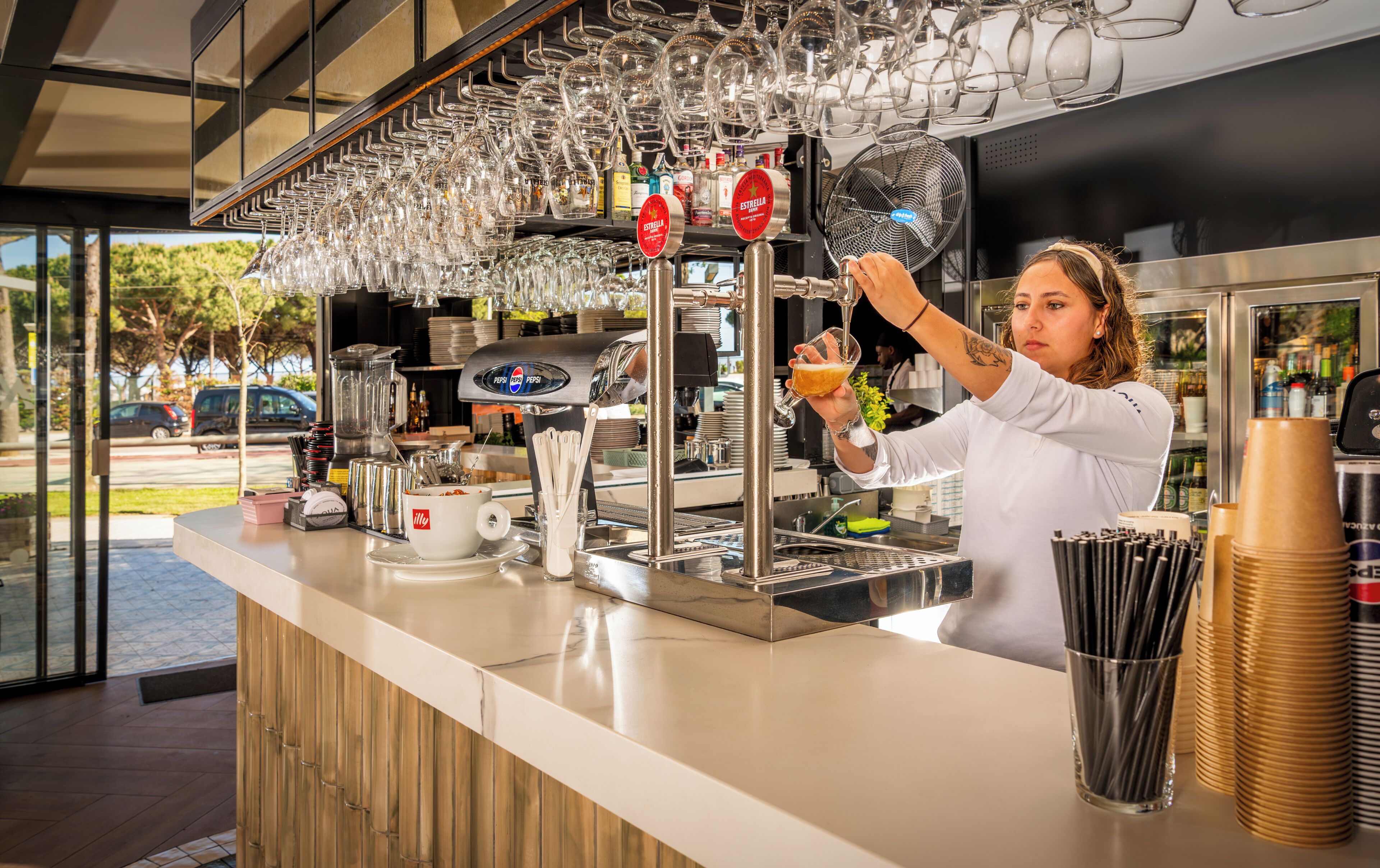 Barista valmistaa juomaa baaritiskillä, jossa on kahvinkeitin, lasiesineitä ja näkymä ulos.