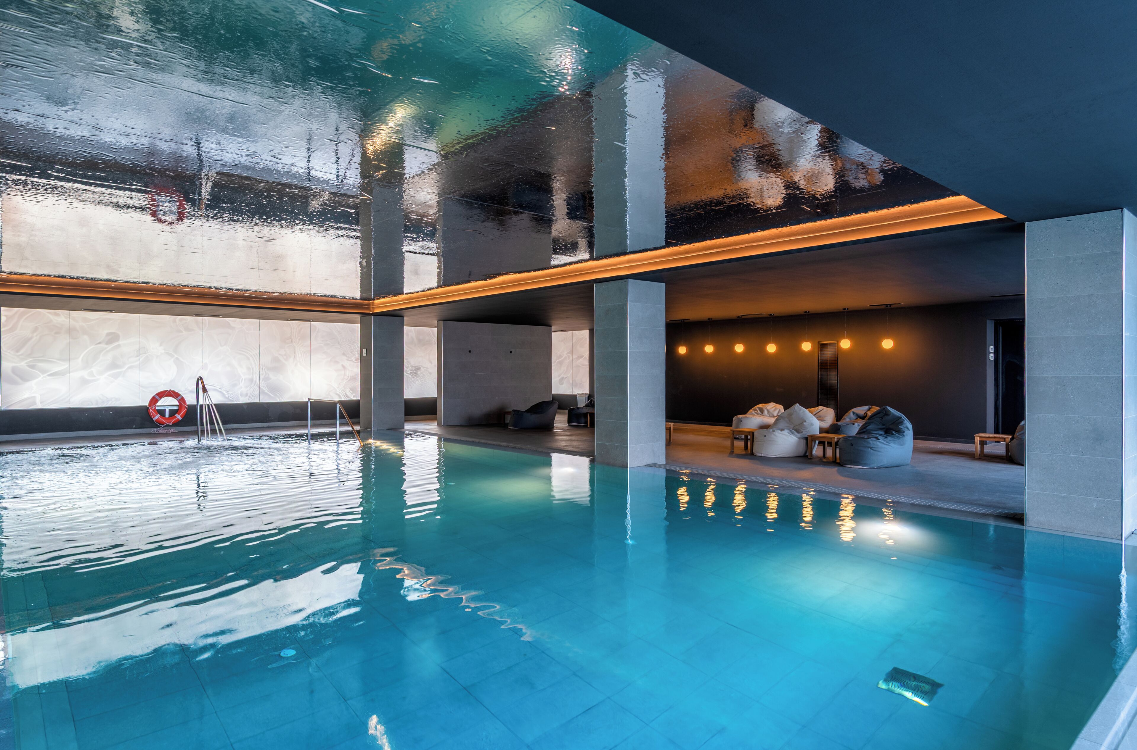 Piscina interior moderna con agua cristalina, pilares de hormigón y zona de estar con pufs y sillas.