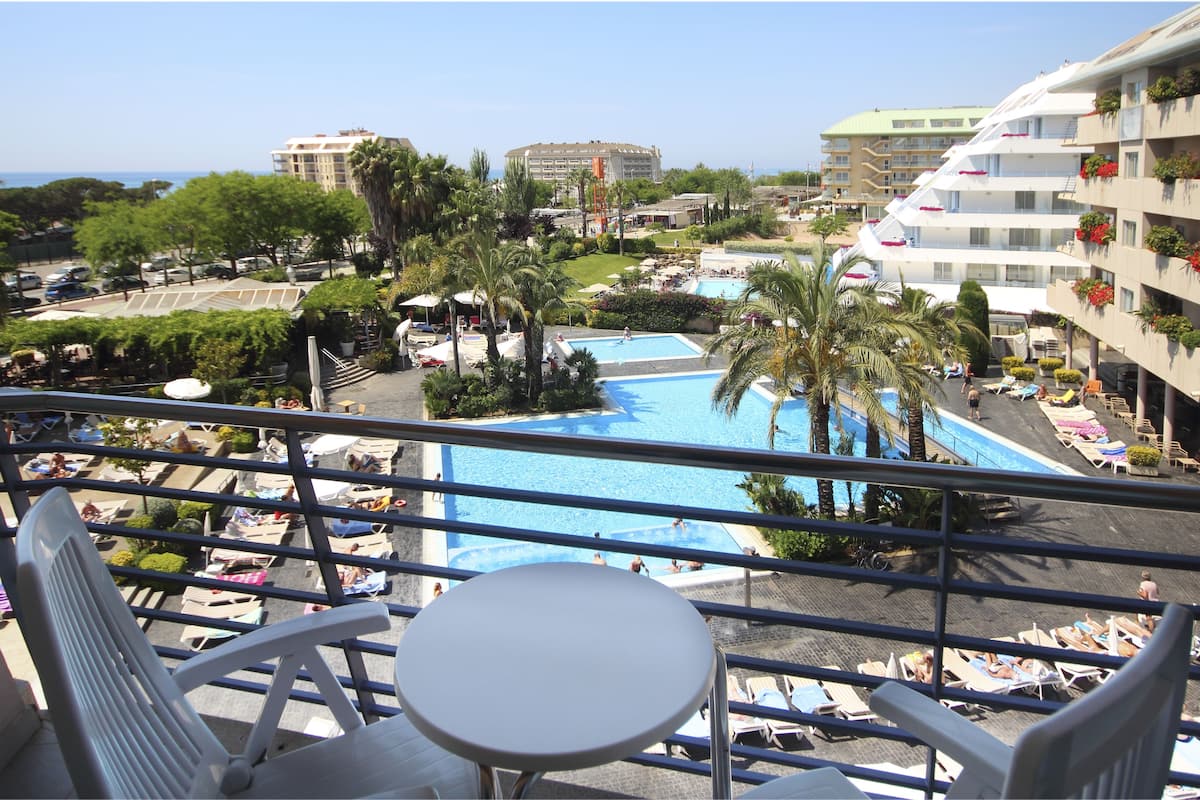 Zona de piscina con sillones de descanso y sombrillas, rodeada de palmeras y edificios.