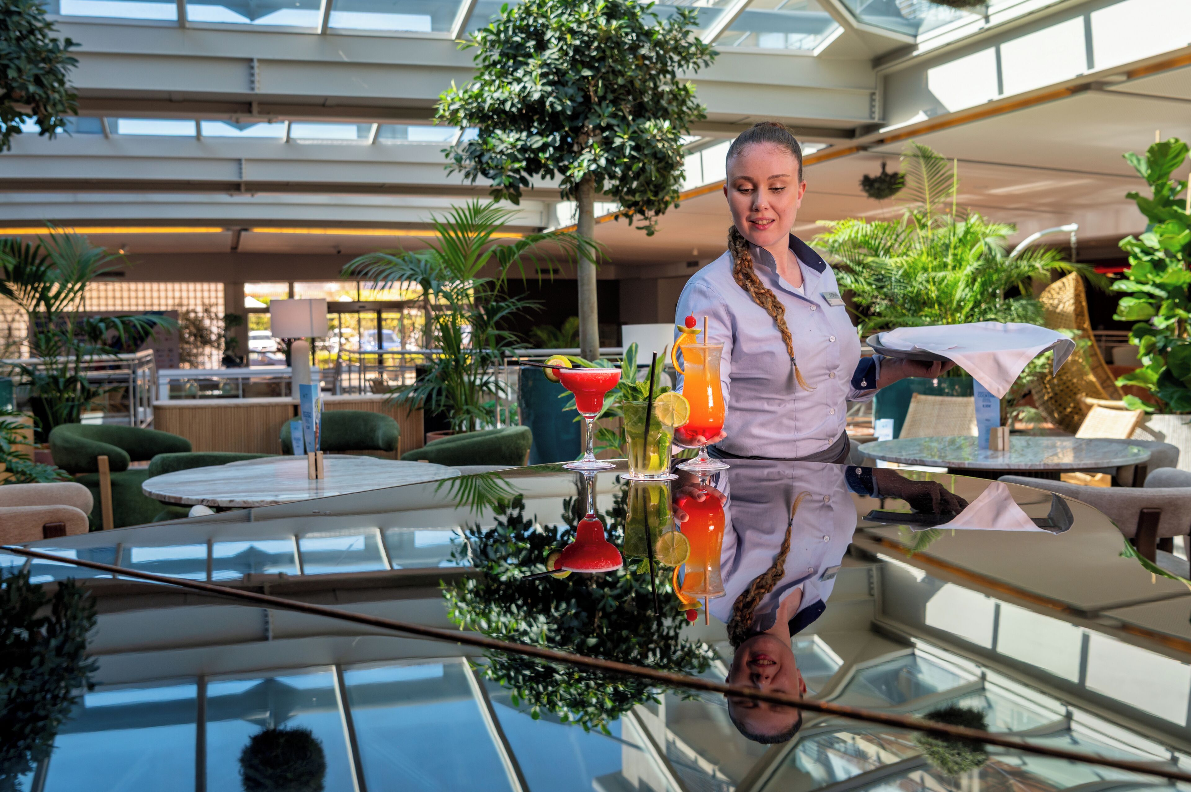 Un hall d’hôtel moderne avec une table en verre qui reflète l’intérieur, un grand arbre et des espaces de détente verdoyants.