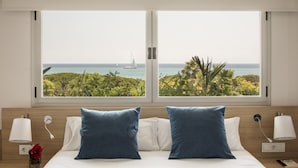 Habitación de hotel con cama, dos almohadas, mesita de noche con lámpara y vistas al océano y un velero a través de amplias ventanas.