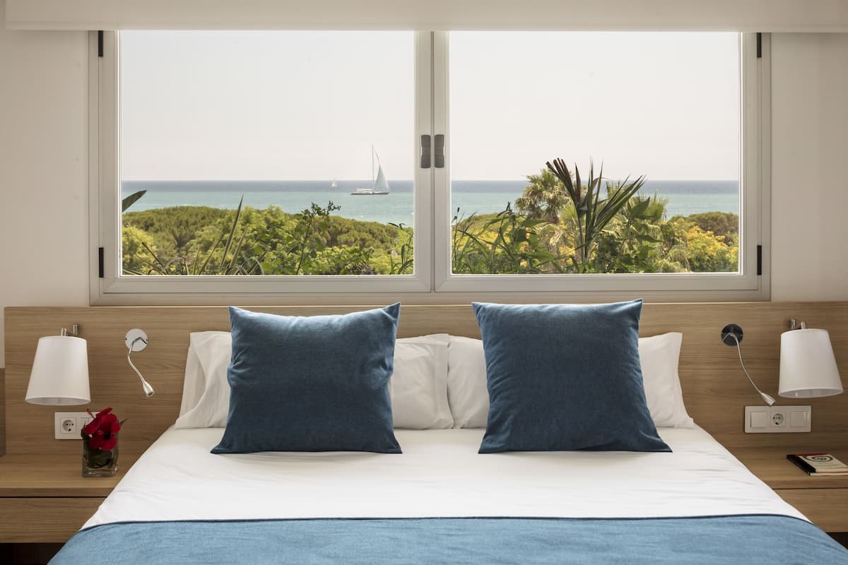 Habitación de hotel con cama, dos almohadas, mesita de noche con lámpara y vistas al océano y un velero a través de amplias ventanas.
