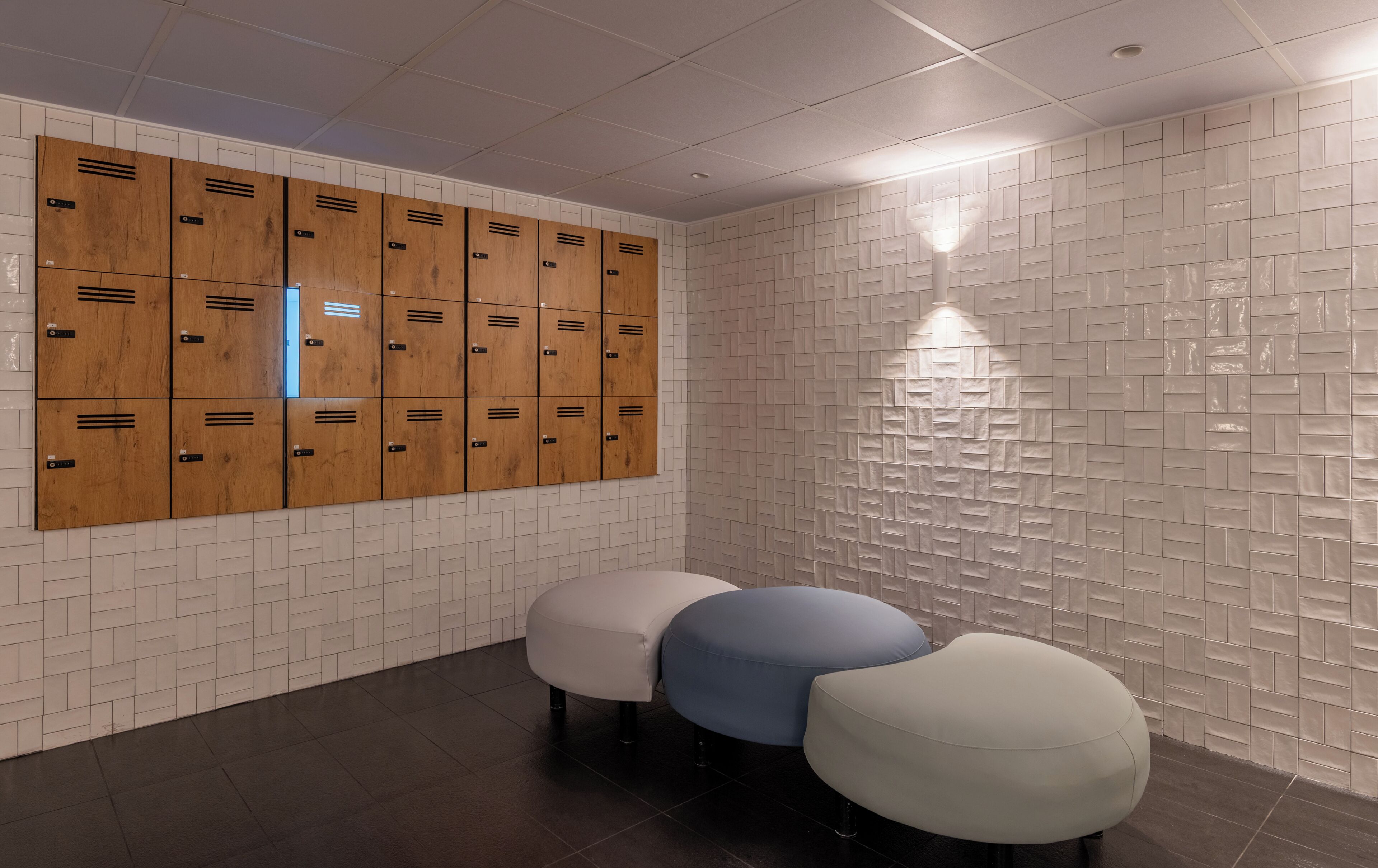 A locker room with wooden lockers, a white tiled wall, and three ottomans.