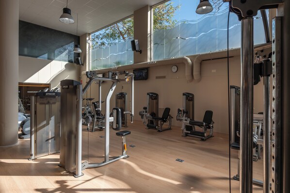 A gym with various exercise machines, large windows, and a high ceiling. - AQUA Hotel Onabrava & Spa (Santa Susanna)