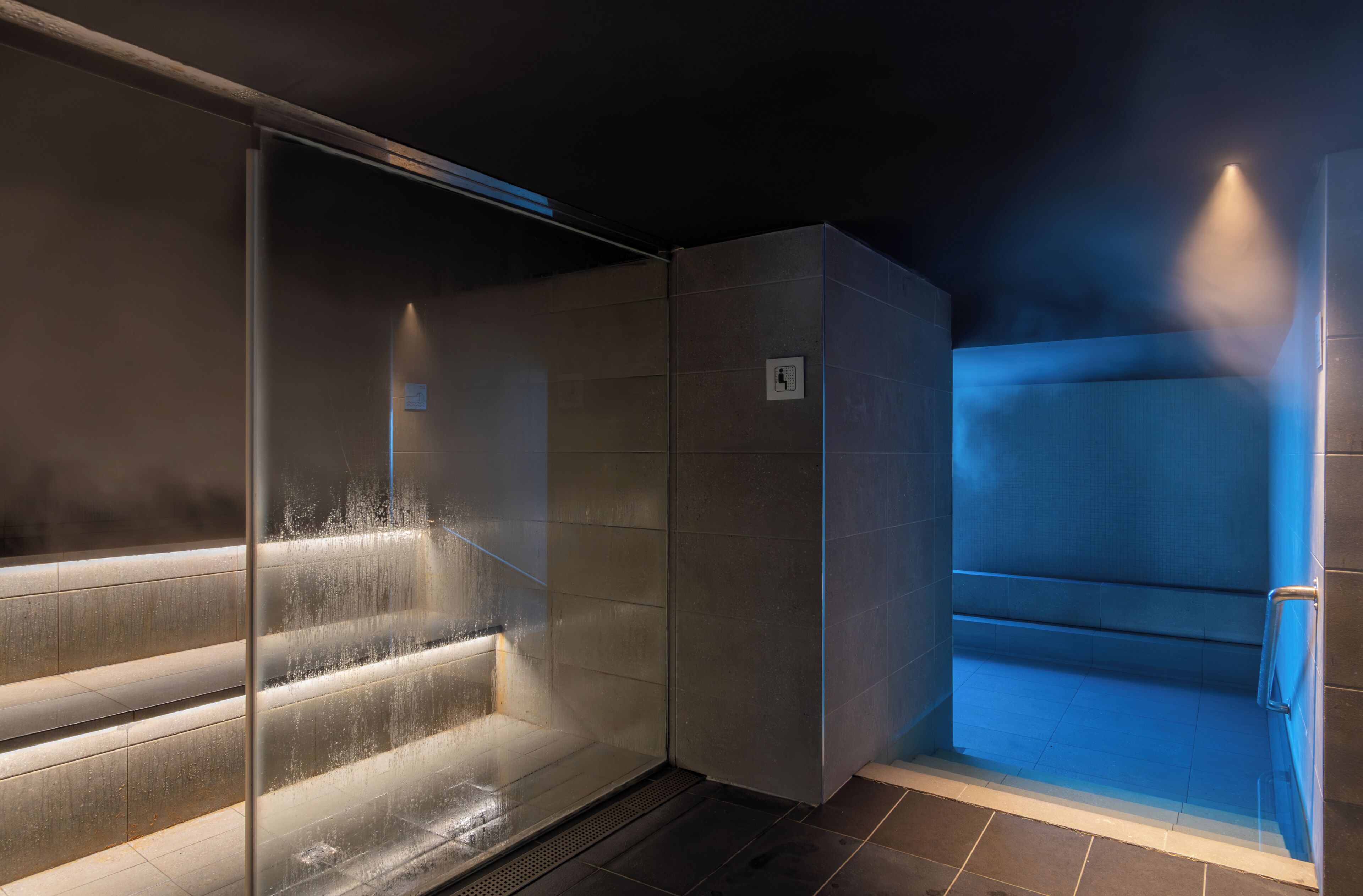 Un área de sauna moderna y cerrada con paredes de vidrio, suelo de baldosas y una esquina iluminada en azul.