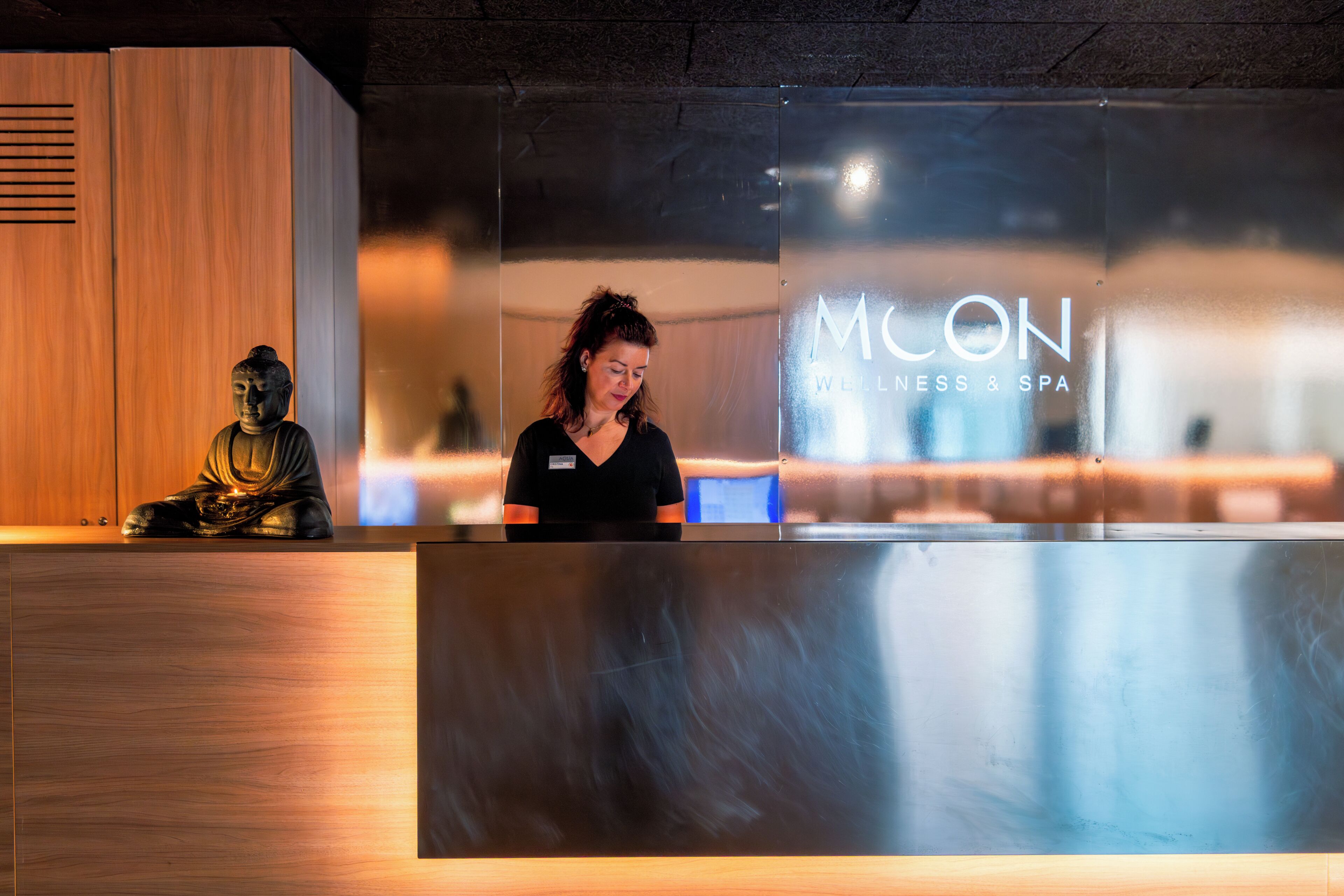Un mostrador de recepción con panel de madera y mostrador metálico, una estatua de Buda y un letrero que dice 'MCON Wellness & Spa'.