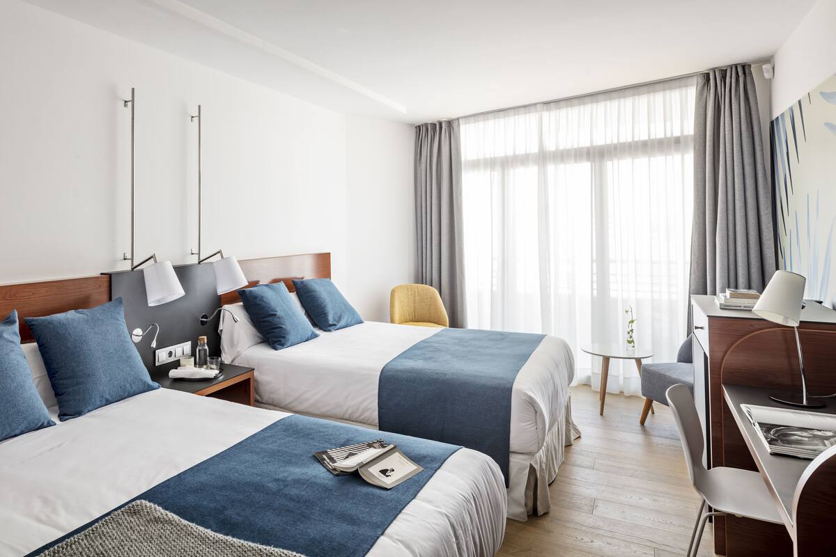 Habitación de hotel con dos camas, un escritorio, una silla y un ventanal con cortinas.