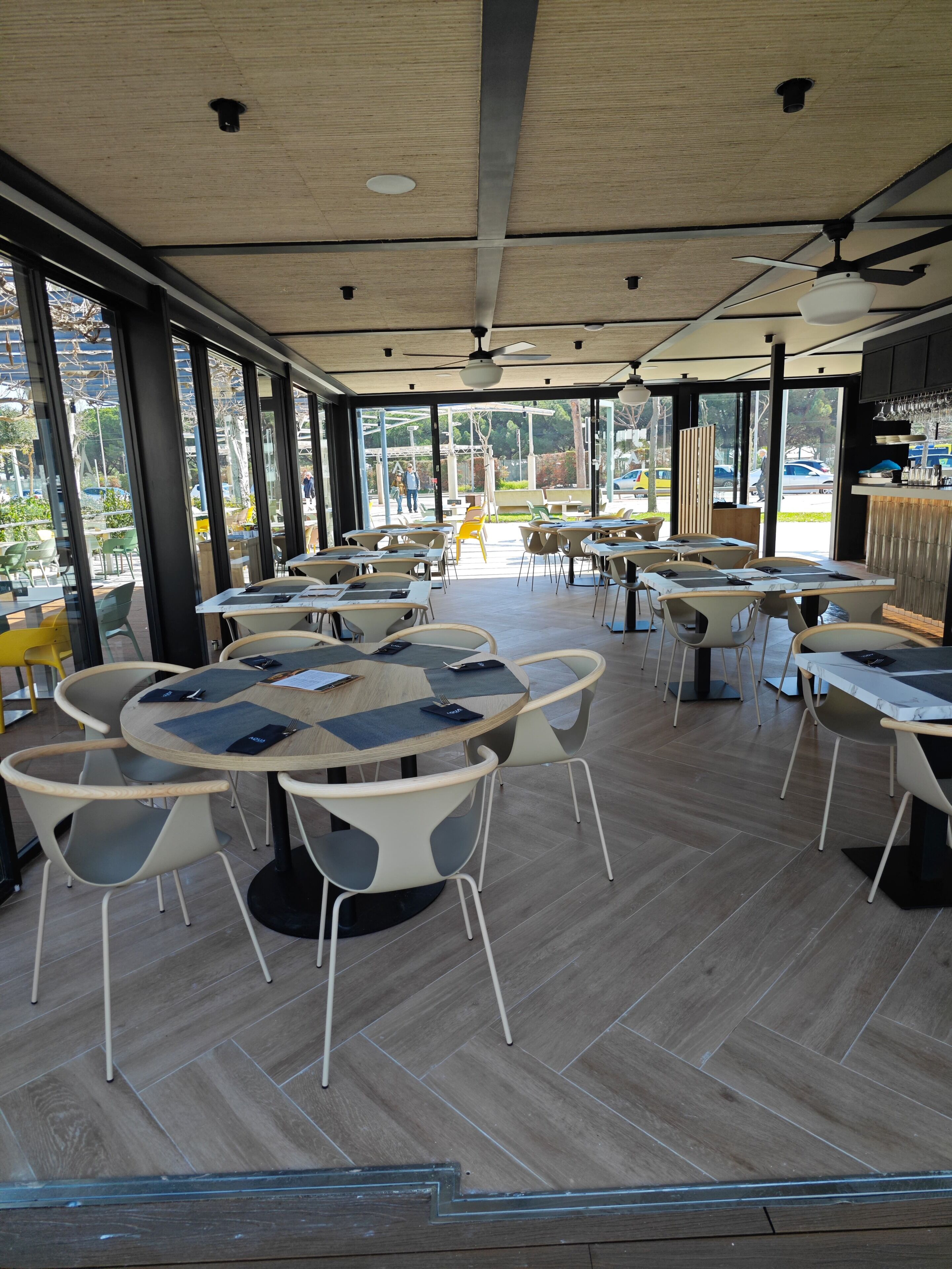 A modern restaurant interior with round tables and chairs, large windows, and a ceiling fan.