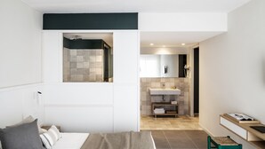 A modern hotel room with a bed, a small stool, a vanity with a mirror, and a shower area. - AQUA Hotel Bertran Park (Lloret de Mar)