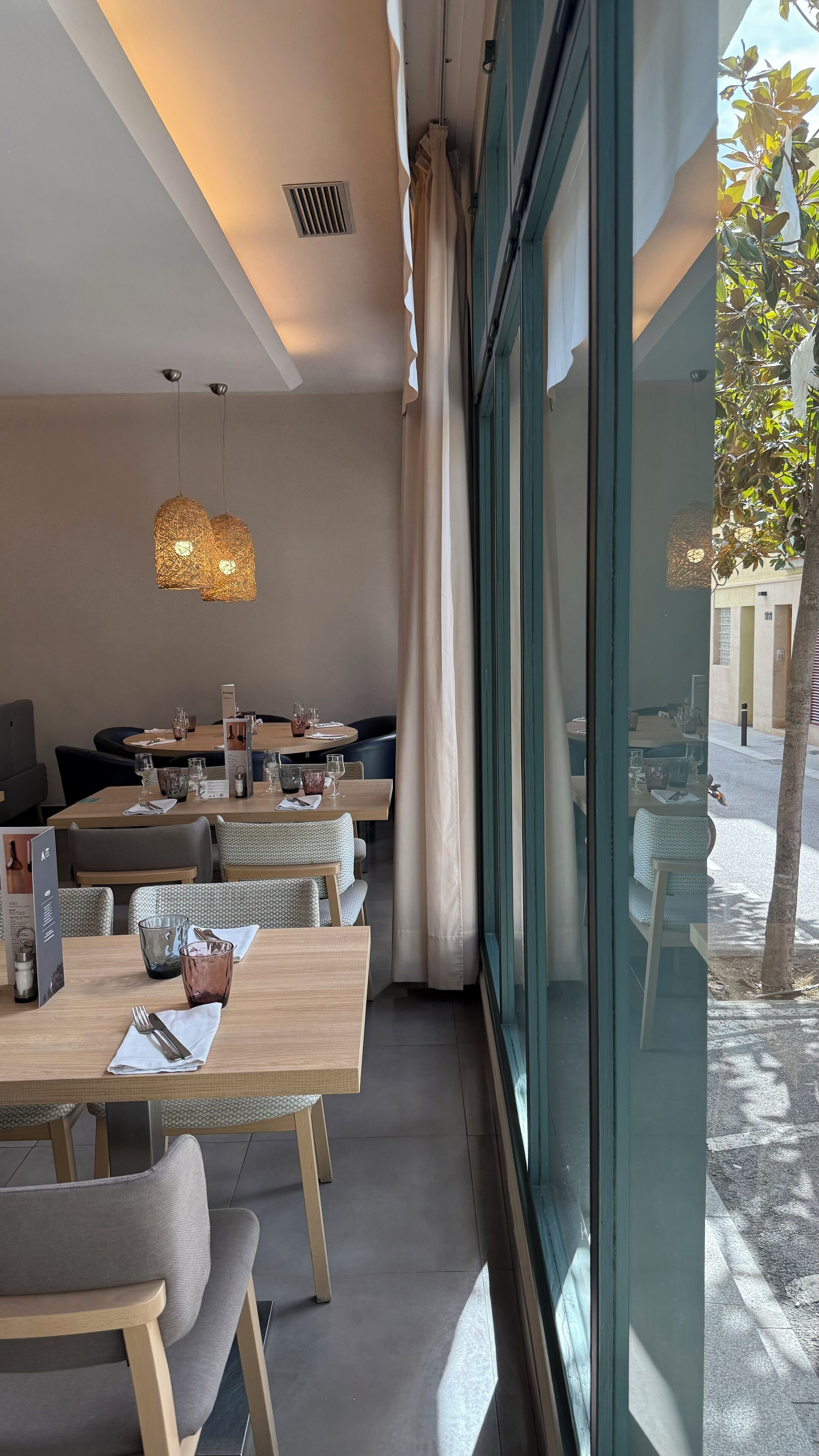 A modern dining area with wooden tables and chairs, hanging pendant lights, and a view of the street through large windows.