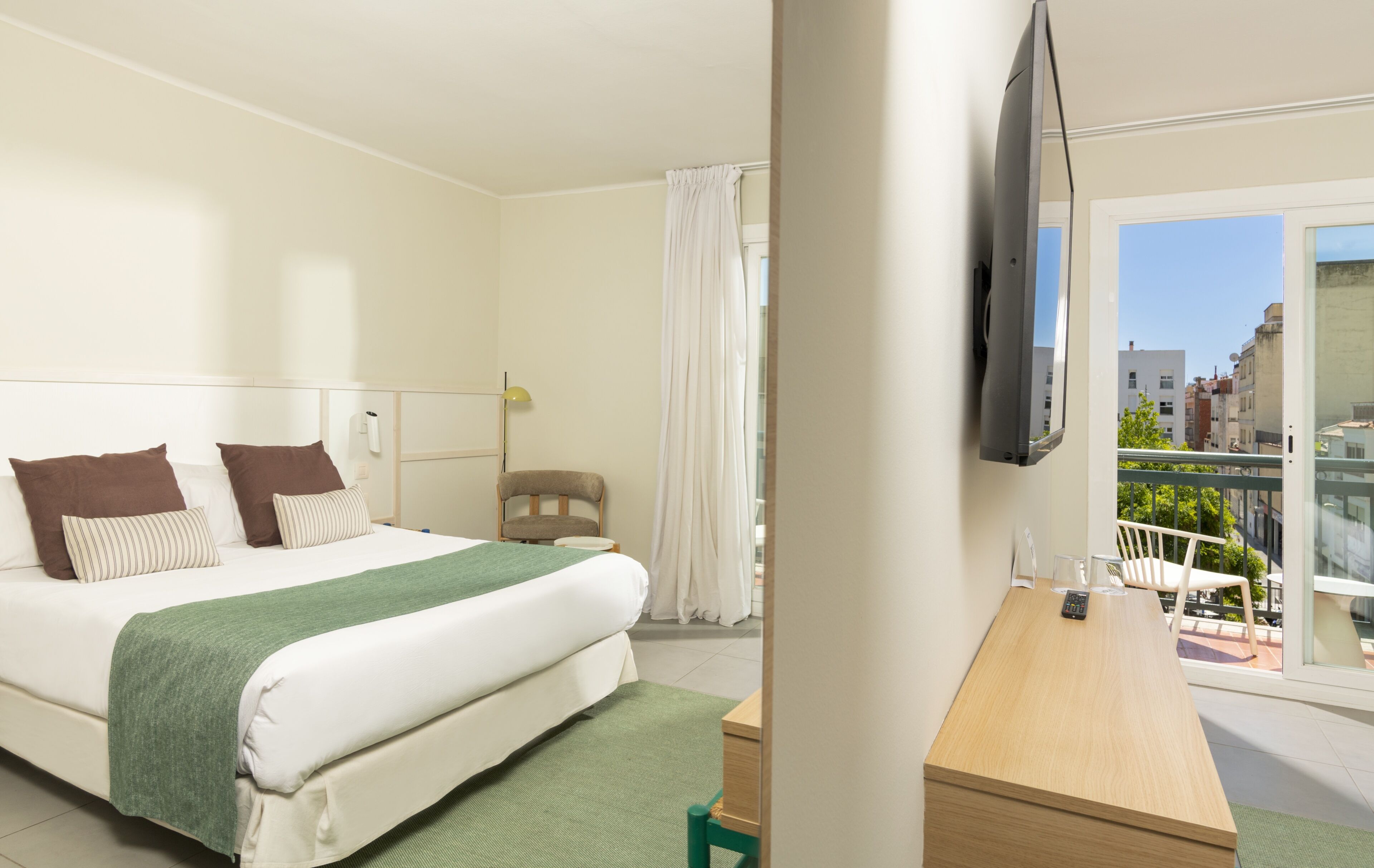 A hotel room with a bed, a desk, a chair, a TV, and a balcony with a view of buildings.