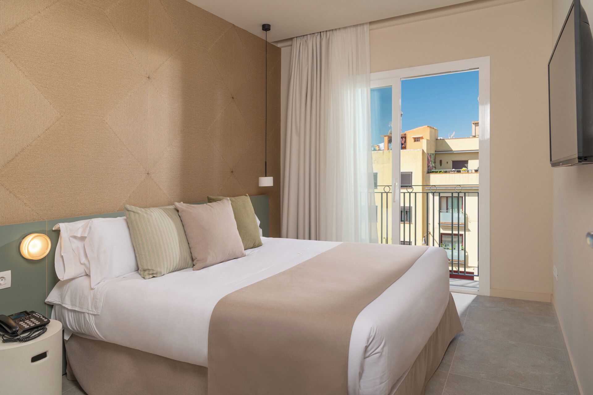 Ein Hotelzimmer mit Bett, Nachttisch mit Telefon, Fenster mit Vorhängen und Blick auf Gebäude.